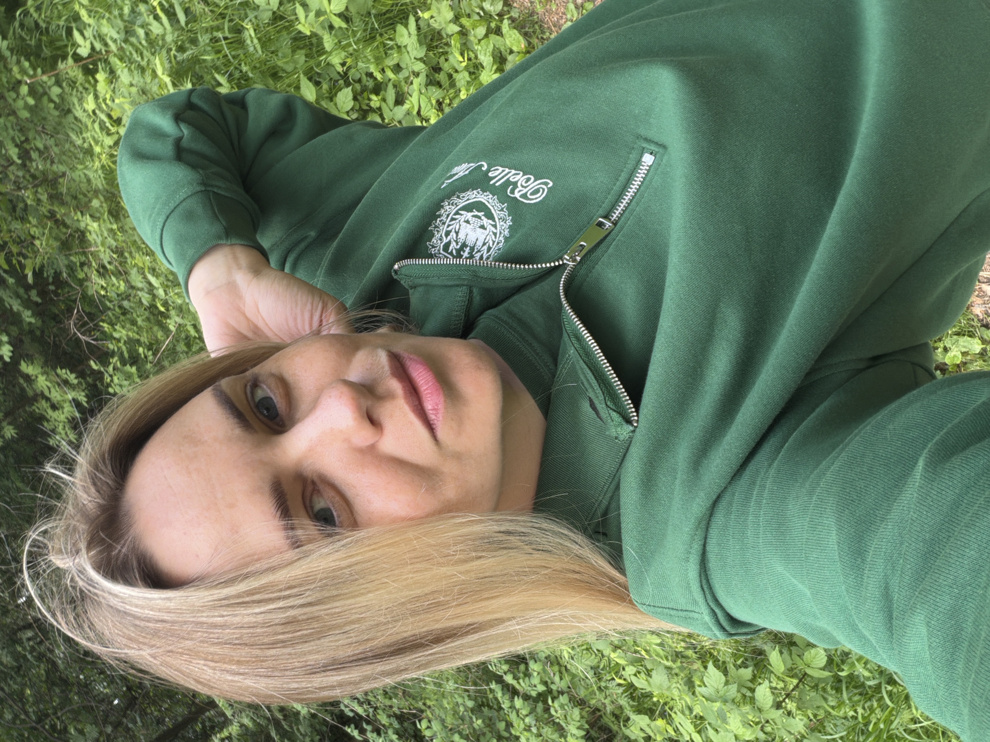 A woman with blonde hair is lying in the grass, likely in a park or garden. She is wearing a green zip-up sweatshirt with a "Belle Vie" logo. The location is unknown, but the prompt suggests Votkinsk, Russia.