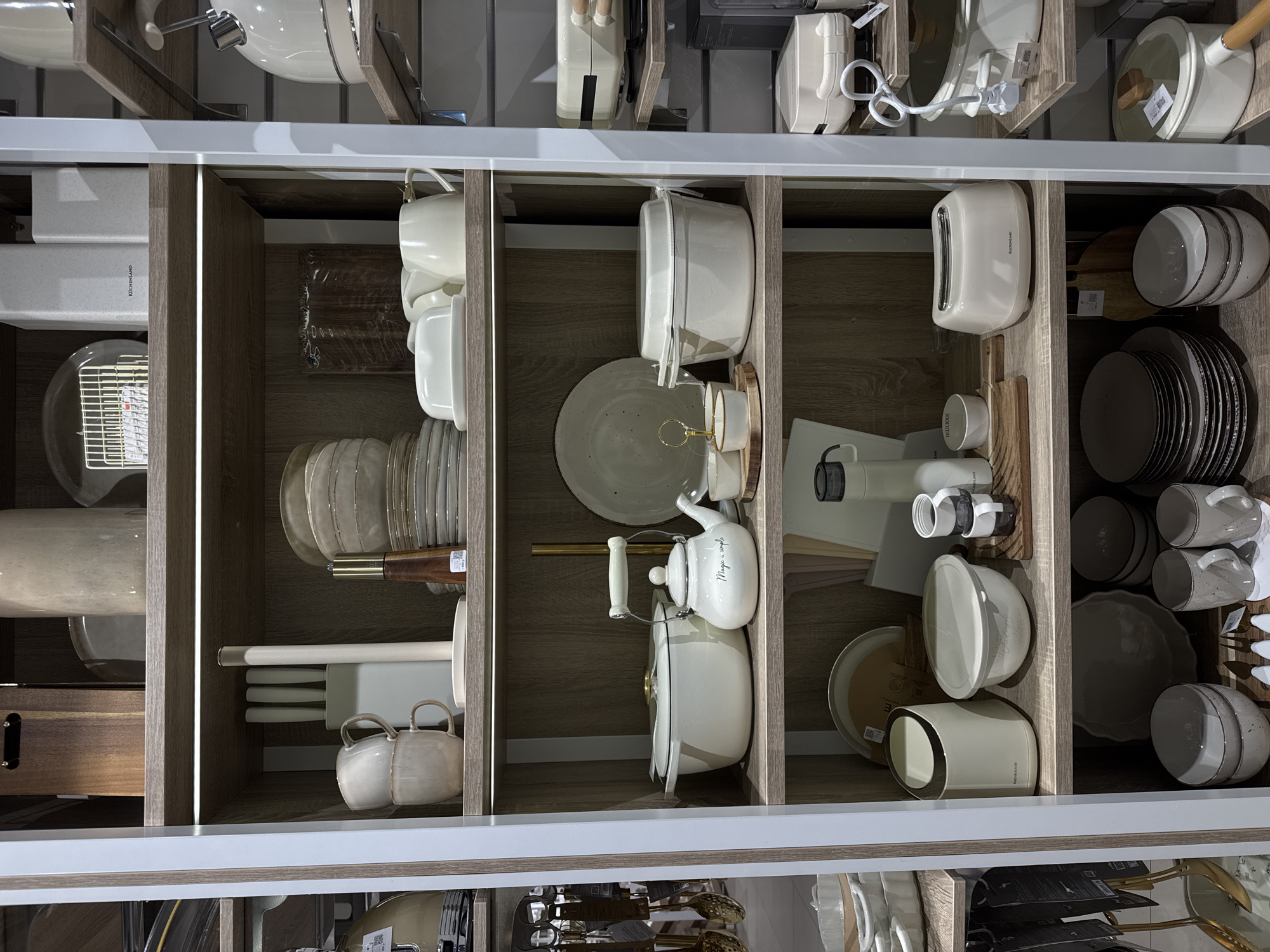 The image shows a display shelf in what appears to be a retail store, likely in Izhevsk, Russia. The shelves contain a variety of kitchenware items, neatly arranged and organized. Items include white and gray dishware such as plates and bowls, cookware such as pots and kettles, and kitchen utensils such as knives and whisks. Some items have labels or price tags attached. The shelves are illuminated by built-in lighting.