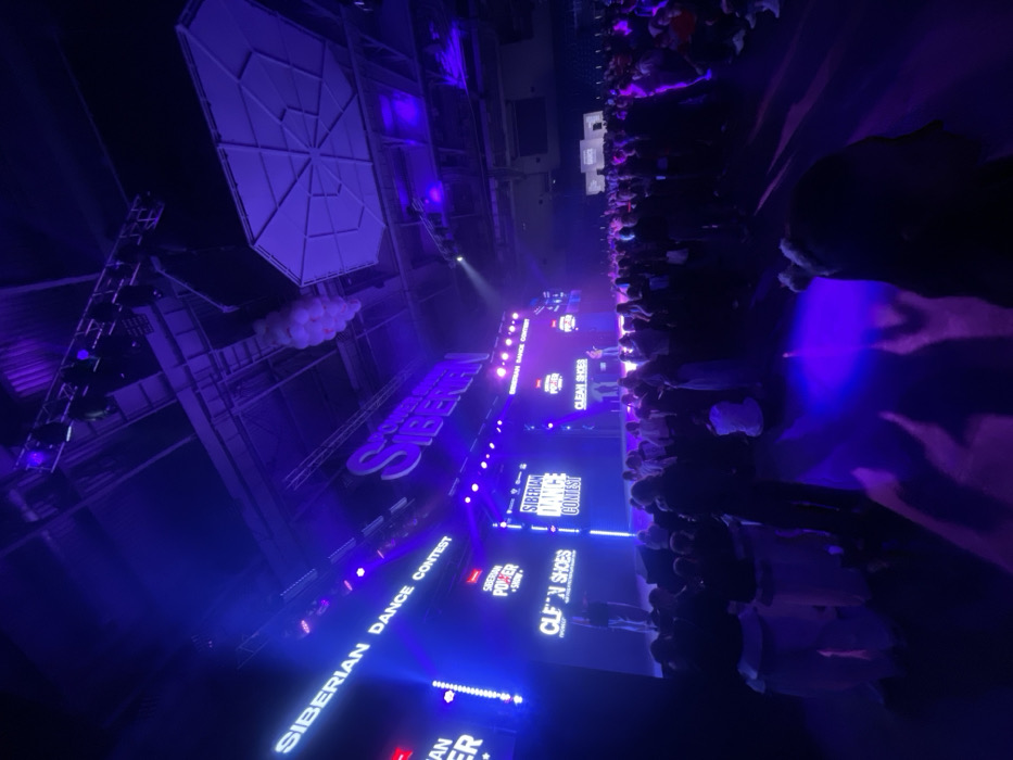 This image depicts a large indoor event taking place in an arena in Krasnoyarsk, Russia, likely the Platinum Arena or a similar large sports and concert complex. The scene is dominated by vibrant purple and blue artificial lighting.
A large stage is visible across the background, featuring multiple prominent LED screens. These screens display text including "SIBERIAN DANCE CONTEST," "SIBERIAN POWER SHOW," and "CLEAN SHOES." A large, stylized "SIBERIAN" logo is suspended above the stage. Two individuals are discernible on the stage, standing or performing.
The high ceiling of the venue reveals exposed structural trusses, lighting rigs, and suspended elements, including a cluster of white balloons or spheres and a large, geometric, light-colored panel.
A substantial crowd of individuals occupies the foreground and midground, standing and facing towards the stage. Their features are largely indistinct due to distance and the low ambient light, but their collective presence indicates a large gathering. One individual's head and shoulders are visible from behind in the immediate foreground. The floor in the foreground reflects the purple stage lighting.