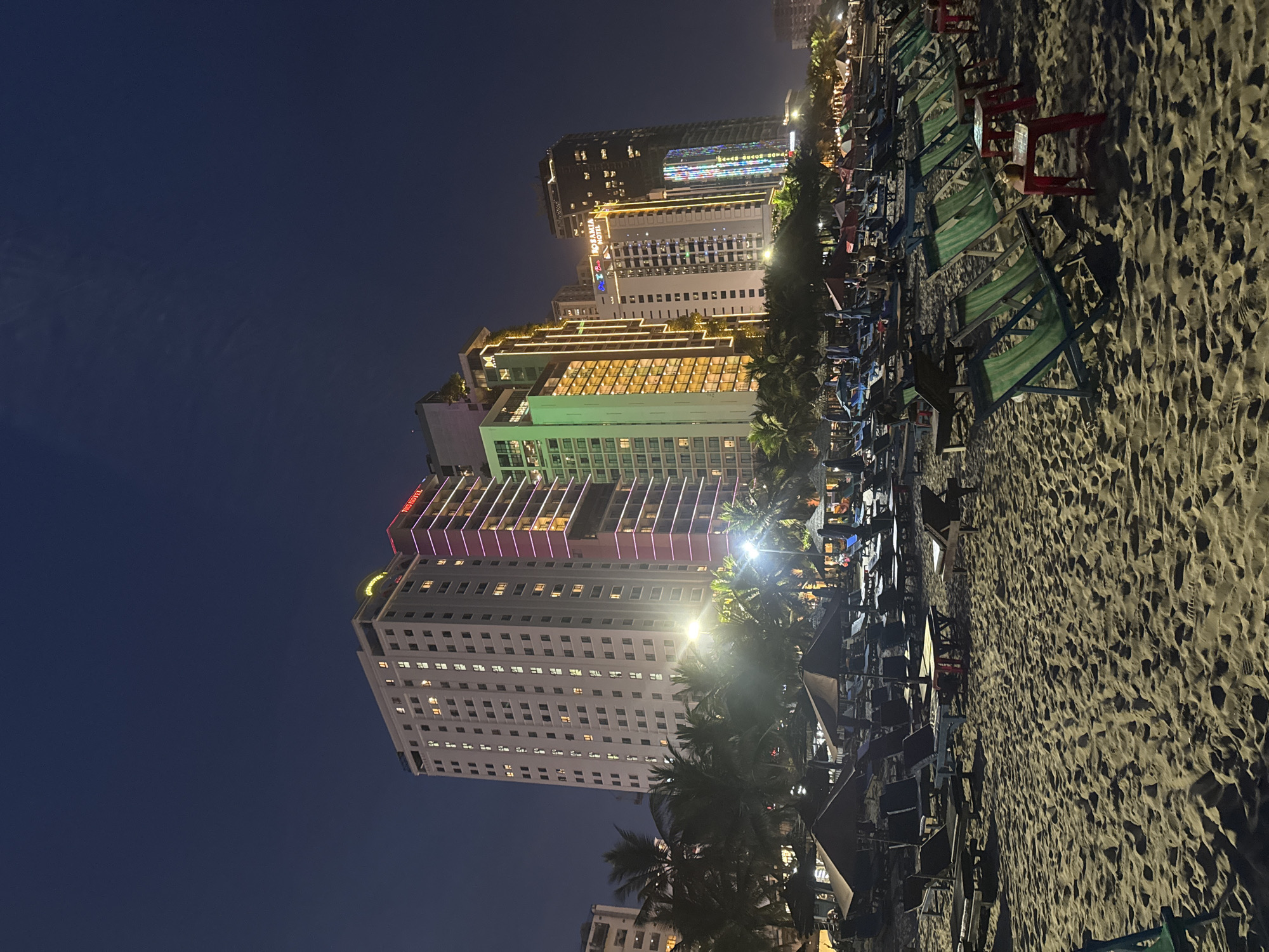 The image shows a nighttime scene on a beach in Đà Nẵng, Vietnam. Several high-rise hotels illuminate the background with their lights. In the foreground, numerous beach chairs are arranged on the sand, seemingly empty. The sky is dark blue. Palm trees are scattered throughout the beach area. The beach is sandy.