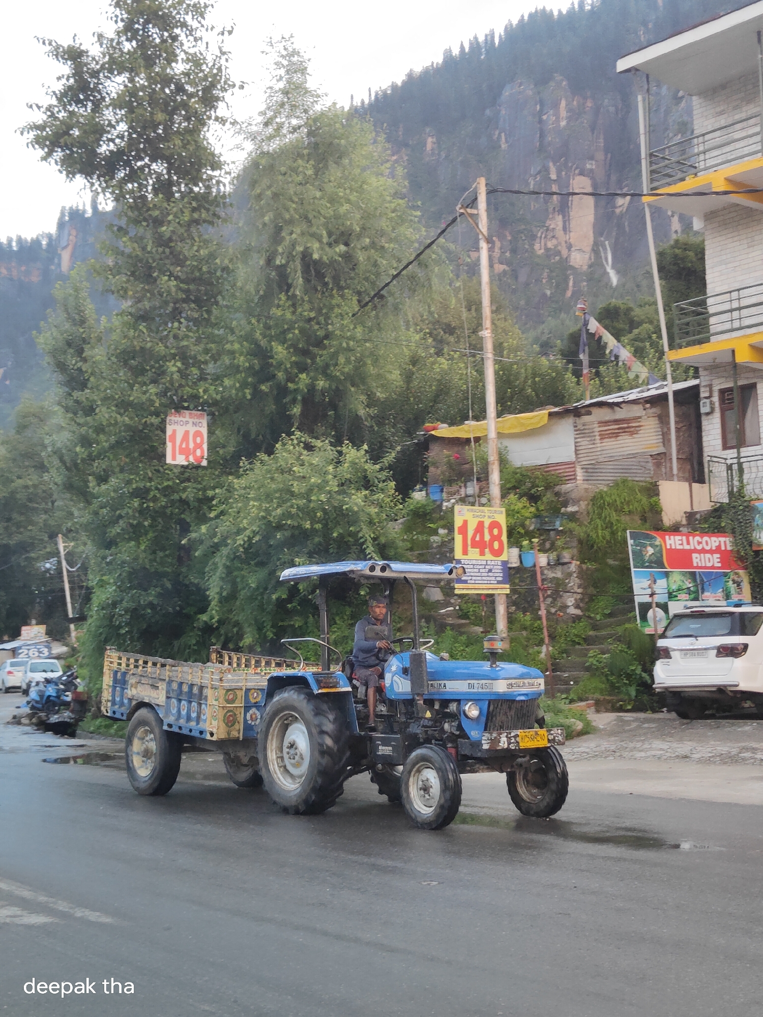 A blue tractor with a wooden trailer is driving down a wet, paved road in Vashisht, India. A man in a dark shirt is driving the tractor. The trailer is filled with what appear to be wooden crates. In the background, there is a steep, rocky mountain covered in trees. A yellow and white building with balconies is visible on the right side of the image. A sign reading "HELICOPTER RIDE" is also visible. A sign with the number "148" is displayed on a tree in the background. The sky is overcast. The words "deepak tha" are visible in the bottom left corner of the image.