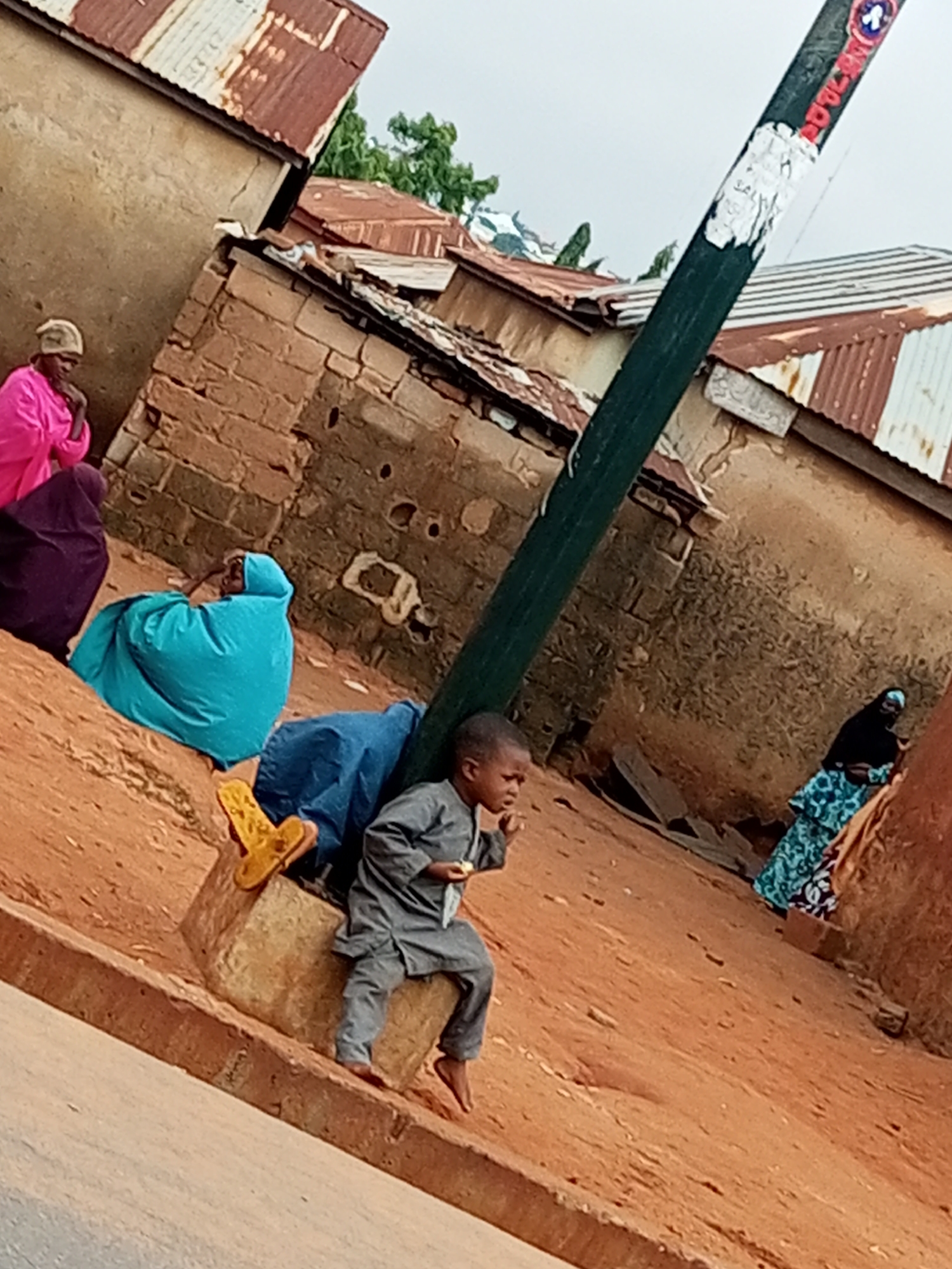 In Tempure, Nigeria, a young boy in a grey tunic and pants sits on a raised concrete ledge, holding a small object in his right hand. He is positioned near a dark green pole that leans diagonally across the frame. Behind him, several people are visible. To the left, three women are seated, one wearing a pink garment and headscarf, another in a dark purple covering, and a third in a bright turquoise headscarf and wrapping. Further to the right, a woman in a patterned blue and black garment stands, wearing a black headscarf. The environment consists of rustic, earth-toned buildings with corrugated metal roofs, indicative of a rural or less developed urban setting. The ground is a reddish-brown dirt. The sky is overcast.