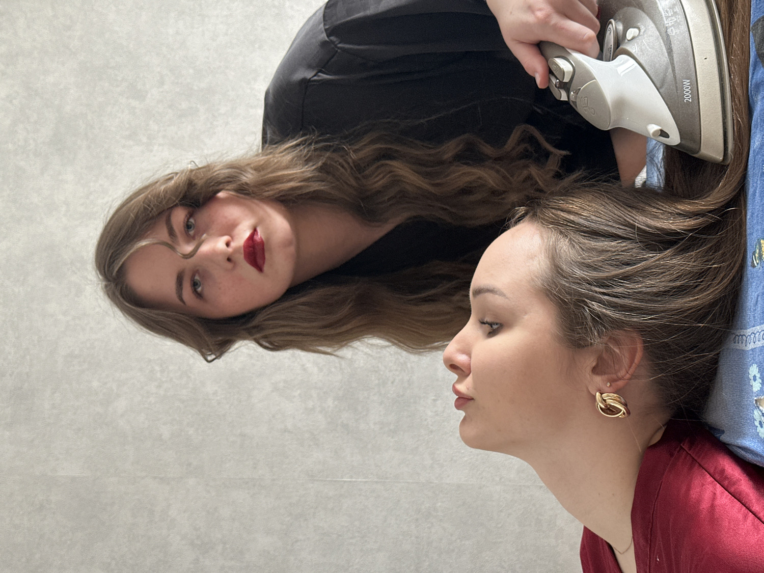 Two young women are depicted in an unconventional indoor scene, likely in Chaikovsky, Russia. The image is oriented vertically, with subjects appearing to lie horizontally against a plain, light grey, textured surface.
In the upper part of the frame, one woman with long, wavy light brown hair looks directly towards the viewer. She has fair skin, light-colored eyes, and is wearing bold red lipstick. Her facial expression is neutral, bordering on alluring. She is dressed in a black top, partially visible.
The second woman is positioned in the lower part of the image, shown in profile, looking to the left. She has straight, dark brown hair that extends towards the right of the frame, appearing to lie flat on a surface. She has fair skin and is wearing a dark red garment and a gold-toned, knotted earring.
A central element of the image is a gray and white clothing iron, held by an unseen hand (belonging to the first woman, implied by proximity) over the hair of the second woman. The text "2000W" is visible on the side of the iron. A blue patterned fabric is seen beneath the iron and adjacent to the second woman's head.
The activity taking place is the seemingly playful "ironing" of the second woman's hair with a clothing iron, creating a surreal and artistic or humorous moment rather than a practical hair-styling session. The plain background offers no specific location cues beyond being an indoor setting. No time of day or weather conditions are discernible.