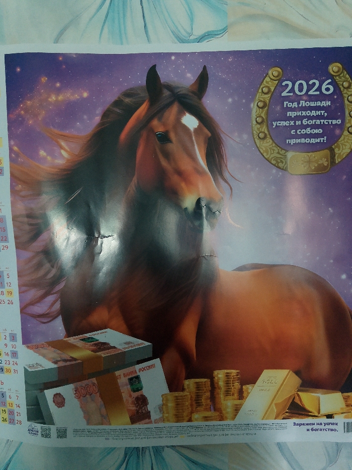 This is a photograph of a calendar page featuring a stylized depiction of a brown horse against a purple starry background. To the right of the horse, a golden horseshoe with the year "2026" and Russian text reading "Year of the Horse brings success and wealth with it!" is prominently displayed. In the foreground, stacks of Russian rubles (5000 denomination) and stacks of gold coins are visible, along with a gold bar. At the bottom, there is a QR code and text in Russian. The overall scene is designed to be auspicious and aspirational, associated with good fortune and wealth. The image does not provide any information about the city of Изыхские Копи, Russia, nor does it contain any people or specific actions.