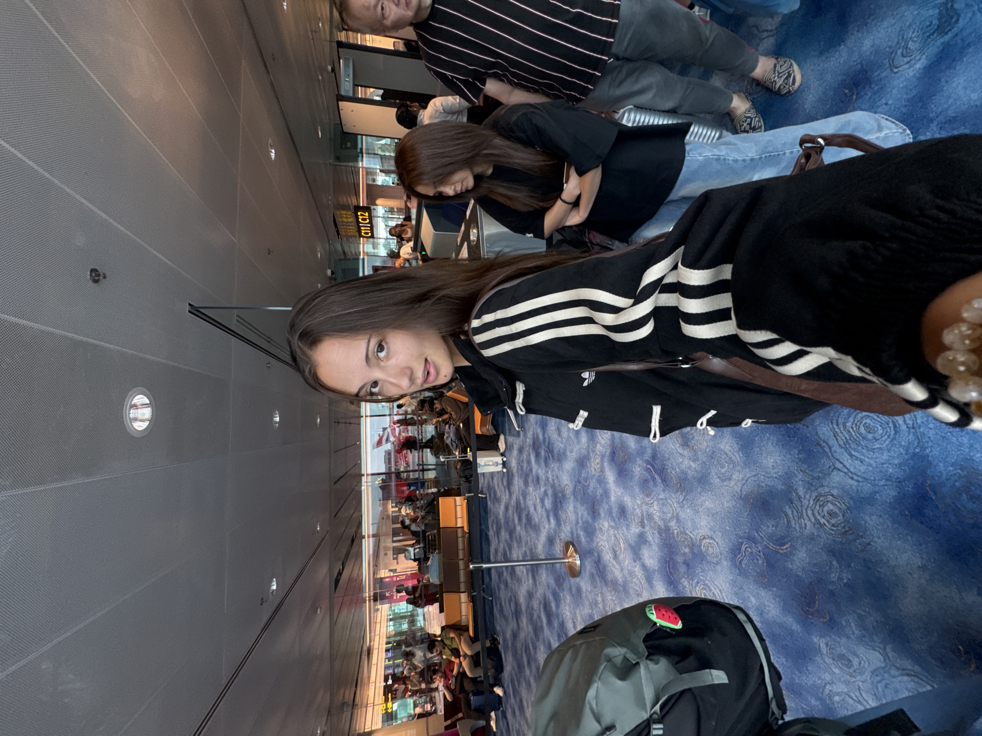 The image shows an airport scene. A young woman in a black and white striped jacket stands in the foreground, looking at the camera. Behind her, a man in a striped shirt and a woman in a black shirt are also present. The background reveals a busy airport terminal with numerous people waiting near gate C11/C12. The airport floor has a distinctive blue patterned carpet. A backpack with a watermelon patch sits nearby. Based on the location of the gate and the visual cues, the airport is likely Singapore Changi Airport in Singapore.