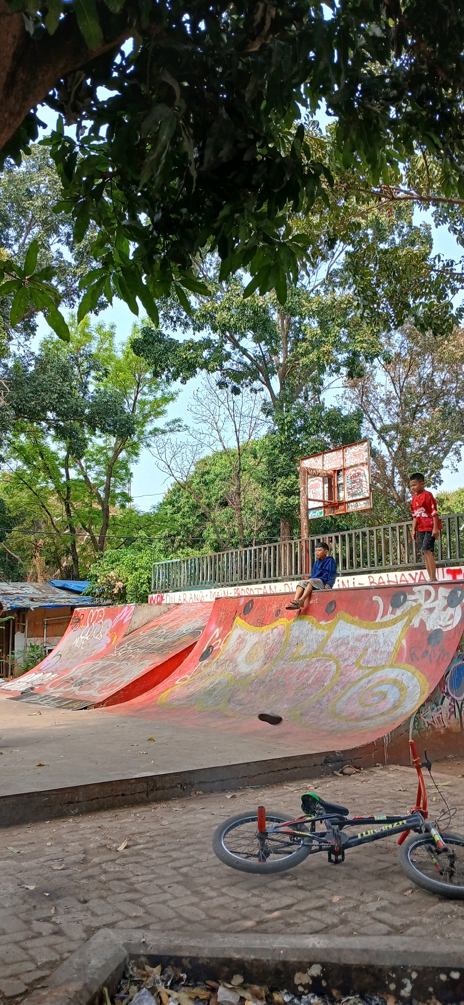 This image depicts a skatepark in the city of Cirebon, Indonesia. In the foreground, a BMX bicycle lies on its side on a paved surface, its frame bearing the inscription "TUFAN ZR". The skatepark itself is characterized by curved ramps painted in vibrant red and yellow, adorned with graffiti.
Two young boys are present in the scene. One, wearing a red shirt with a white design, stands near a rusty basketball hoop, seemingly observing. The other boy, dressed in a blue long-sleeved shirt and khaki shorts, sits on the edge of a ramp, his feet resting on the lower part. Behind the skatepark, a metal fence borders the area, and beyond that, trees and vegetation create a natural backdrop. A small, makeshift structure with a blue tarp roof is visible to the left. The ground is covered with leaves and debris, suggesting an outdoor environment. The overall impression is of a recreational area, likely a local community space.