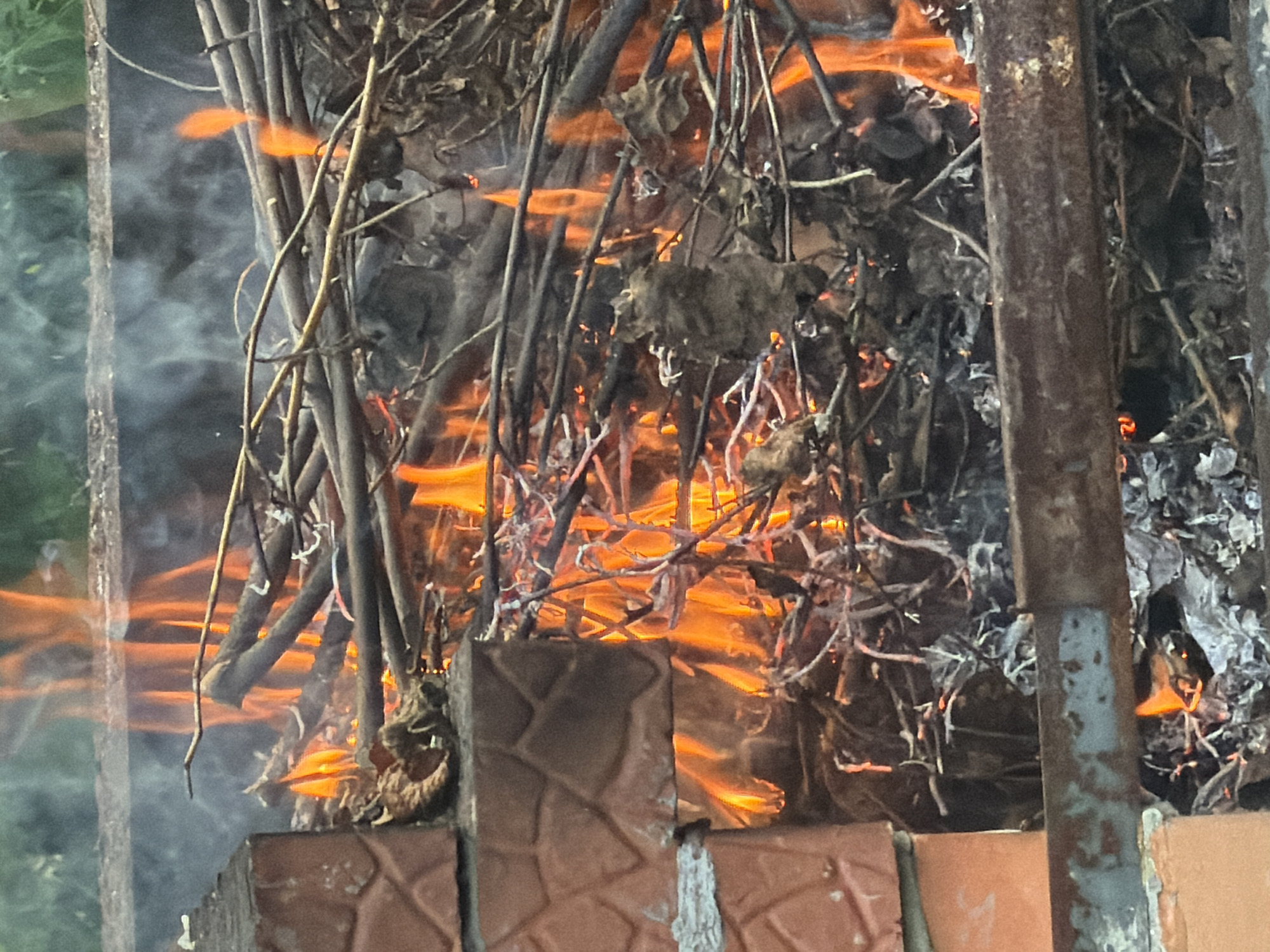 A close-up shot captures a bonfire in a fire pit. The fire pit is constructed with red brick on the left and a rusty metal bar across the front. Dry branches and leaves are actively burning, with visible flames in orange and yellow hues. Smoke rises from the fire. The background is blurred, showing hints of green foliage. The scene does not provide any specific information that would allow for localization to the city of Yekaterinburg, Russia.