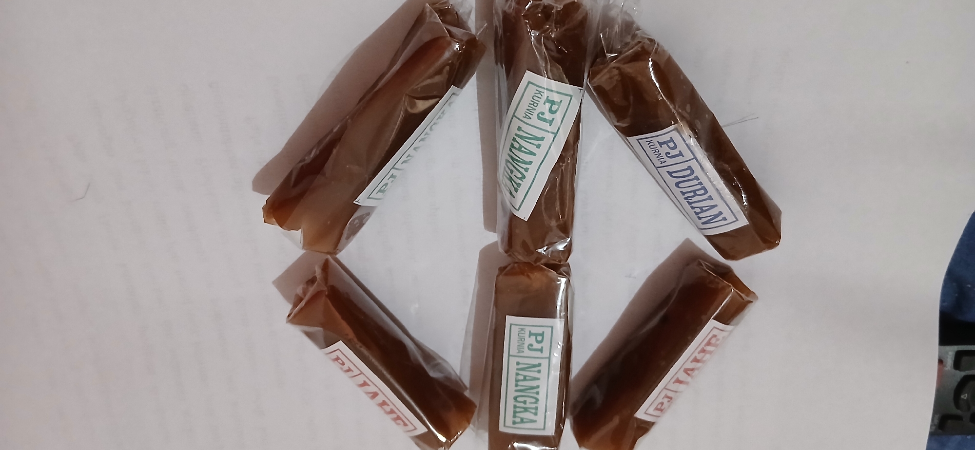 In this close-up, overhead shot, six individually wrapped confectioneries are arranged on a white surface, likely paper, with some printed text visible underneath. The confectioneries are uniformly brown, elongated, and cylindrical, with slight variations in their shape due to the wrapping. Each is enclosed in clear plastic packaging.
Five of the six items have visible labels. Three of the labels clearly read "PJ NANGKA" with "KURNIA" written below in a smaller font. One of these "PJ NANGKA" items is positioned centrally and vertically, while another is to its right, angled slightly. A third "PJ NANGKA" is visible on the left side, angled upwards.
Another confectionery, positioned to the lower left, has a label that reads "PJ DURIAN" with "KURNIA" below.
A sixth confectionery, located in the upper left of the frame, has a partially visible label that reads "PJ TAHF". The top edge of this label is visible, showing red lettering on a white background.
The background is a plain white surface, likely paper, with faint printed text that is mostly out of focus. The overall lighting is diffused, casting soft shadows from the confectioneries. To the far left of the image, a dark blue object with some buttons, possibly a remote control, is partially visible. The scene is static and appears to be a product display or a casual arrangement of food items. There is no indication of a specific location within the city of Samarinda, Indonesia, beyond the potential origin of the product as suggested by the labels.