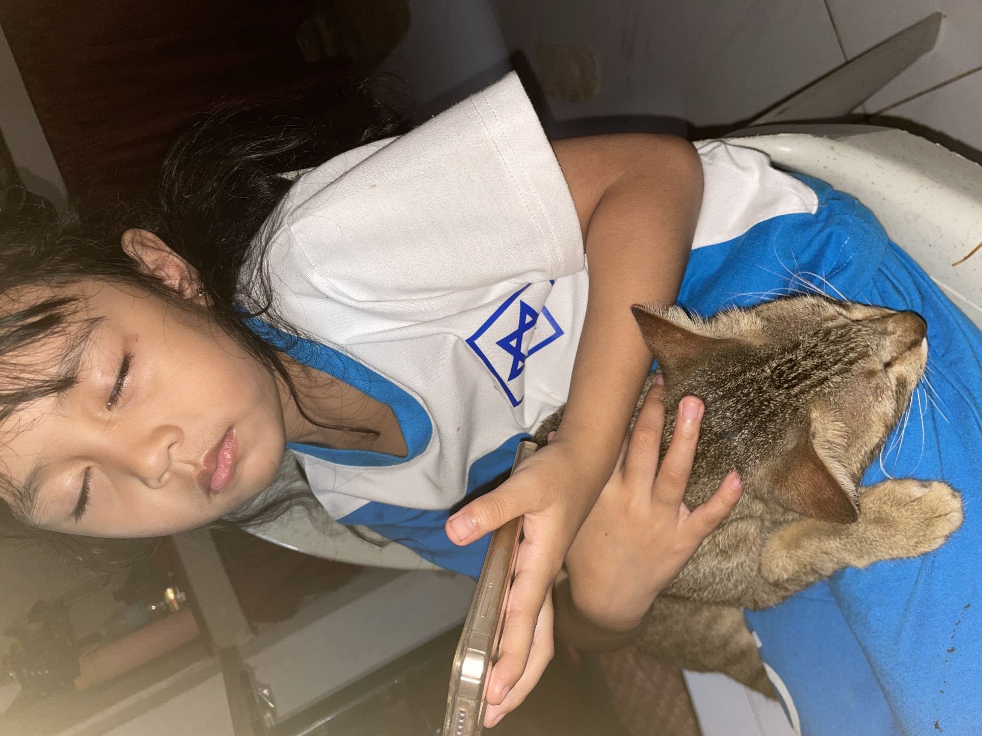 A young Asian girl with her eyes closed is sitting and holding a tabby cat. She is wearing a blue and white t-shirt with a geometric logo on it and blue pants. Her left hand is holding a smartphone. The cat is lying on her lap, with its head resting on her arm. The background appears to be an indoor setting with some furniture visible. The lighting suggests it is nighttime or a dimly lit room. The overall mood is peaceful and intimate. The image does not contain any specific visual cues that would allow for identification of its precise location within Davao City, Philippines.