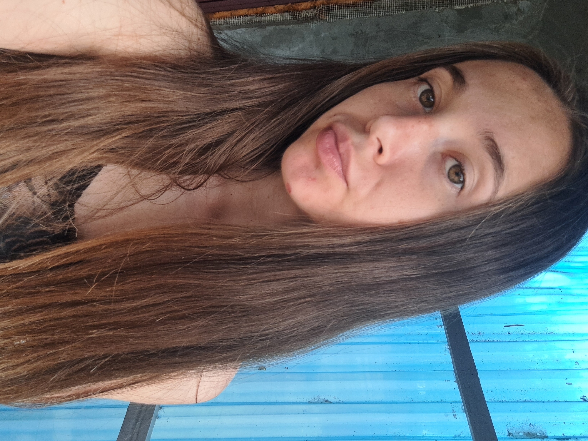 A young woman with long, brown hair is photographed from the chest up, in what appears to be an outdoor setting. She is facing the camera with a neutral expression. Her skin is fair with some freckles and a small blemish on her chin. She is wearing a dark, lacy top. In the background, a blue corrugated metal wall is visible, along with a portion of a gray, rough-textured wall. There is also a dark wooden frame with mesh on the left side of the image. The lighting suggests it is daytime. The presence of the blue corrugated metal suggests a utility area or outbuilding, common in various residential or industrial settings. The overall scene is simple and does not contain any specific indicators of the city of Vypasne, Ukraine.