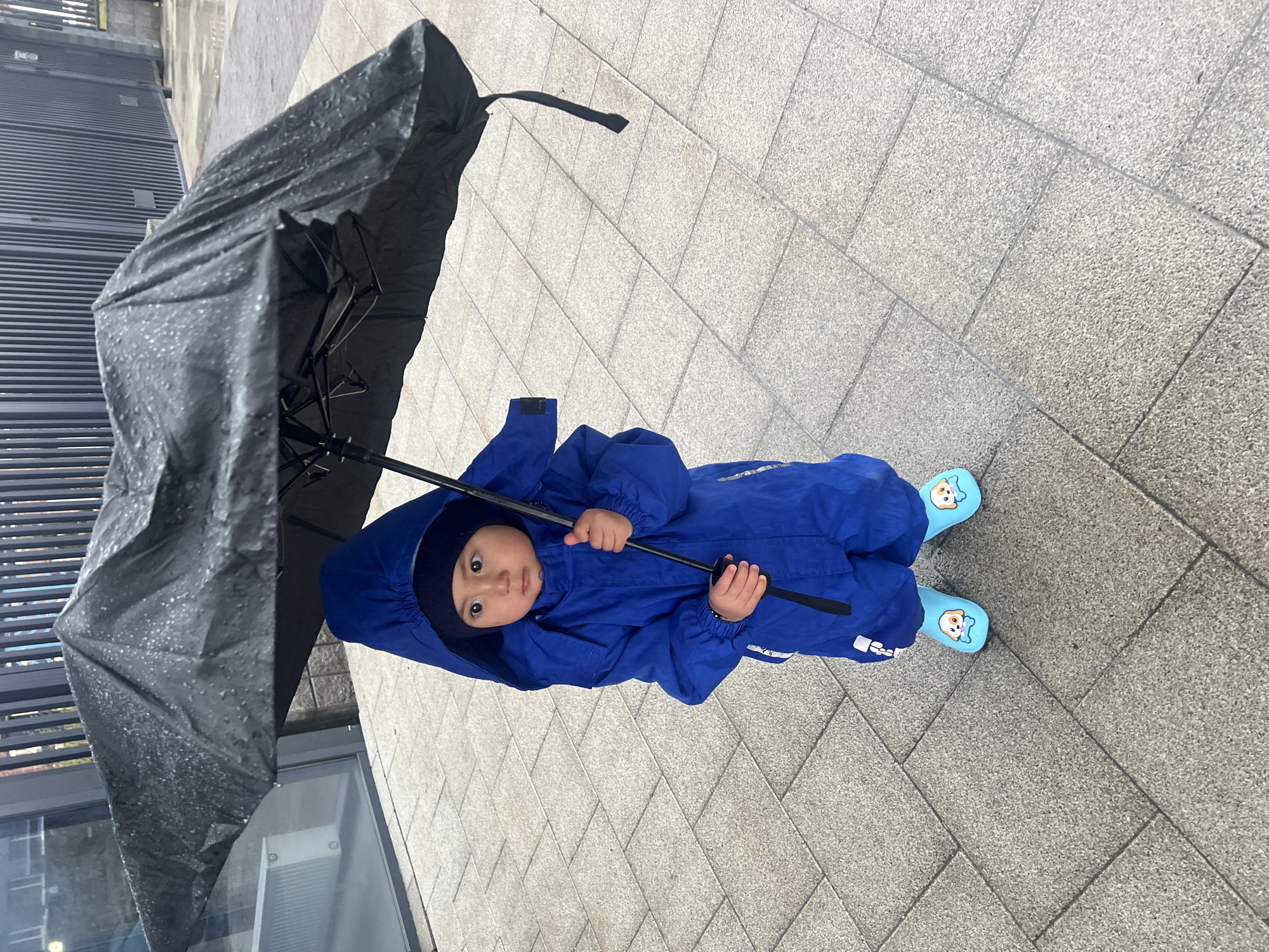 A young child, likely a toddler, is depicted standing outdoors on a paved surface in Tyumen, Russia, holding a large black umbrella. The child is dressed in a bright royal blue full-body waterproof rainsuit with a hood, covering their head. Their light blue boots feature a distinct cartoon dog face design.
The black umbrella, held with both hands by the child, is visibly covered in numerous water droplets, indicating that it is raining or has recently rained. The child's face is visible within the hood, looking directly at the camera with wide, dark, and seemingly curious eyes. Their expression appears neutral and attentive.
The setting includes a light gray, textured paved ground composed of square and rectangular tiles. In the background to the left, a modern building with a dark, horizontally-ridged facade is partially visible. The scene suggests a daytime outdoor activity during wet weather. A small, dark patch, possibly a label or logo, is visible on the left sleeve of the child's rainsuit, and a faint reflective pattern on the right leg, though no legible text can be identified.