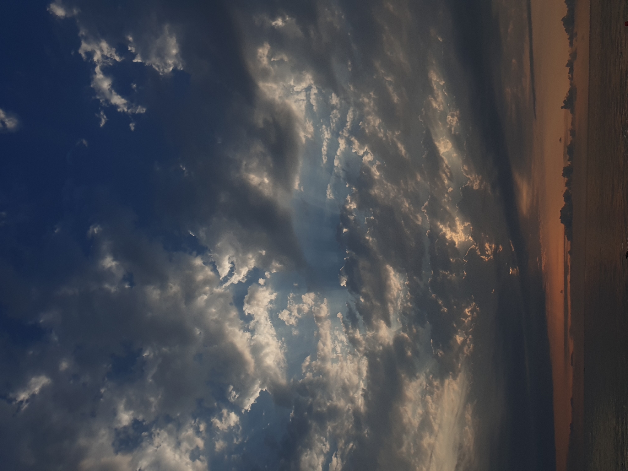 A dramatic, overcast sky looms above a calm body of water, likely the Black Sea, at the edge of Batumi, Georgia. The clouds are a mix of dark grays and whites, with shafts of sunlight breaking through in the upper center of the frame, creating crepuscular rays. The sun itself is not visible, but its light illuminates patches of the clouds from behind. The horizon line is low, with a thin strip of reddish-orange light suggesting a sunset or sunrise. The water is smooth and dark, reflecting the muted colors of the sky. There are no visible people or man-made structures in the immediate foreground or on the water. The scene conveys a sense of atmospheric intensity and natural beauty.