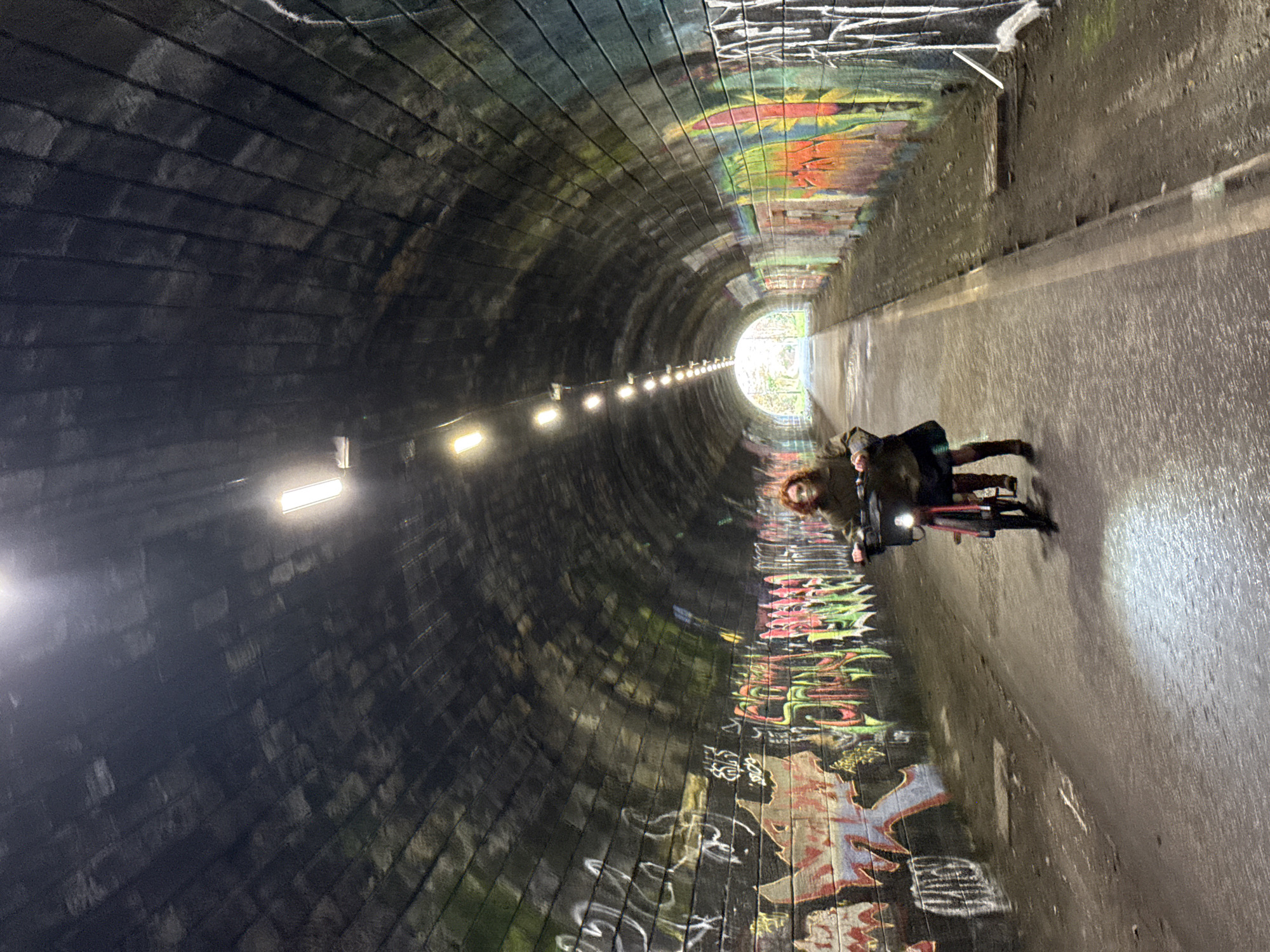 A long, arched tunnel with brick walls receding into the distance with graffiti along its length is illuminated by overhead lights. A person is riding a bicycle toward the end of the tunnel where light is visible. The scene is possibly located in the city of Unknown, Unknown.
