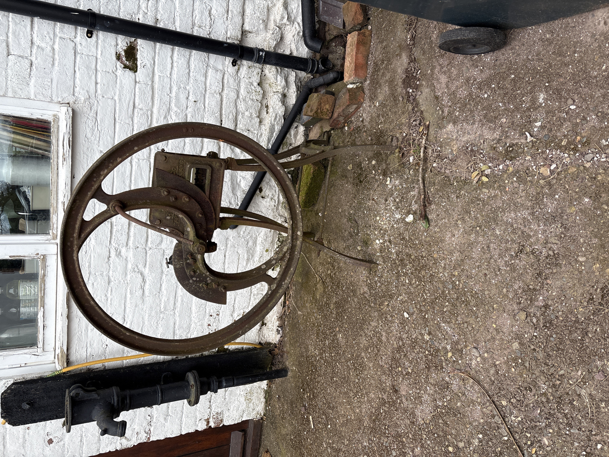 The image, oriented horizontally, features a vintage chaff cutter positioned against a white brick wall. The chaff cutter is made of rusted metal and has a large, circular wheel as its primary feature. The mechanism of the cutter includes blades and a feed trough.
The wall is part of a building exterior, marked with signs of age and weathering. A window frame, painted white, is visible to the left of the cutter. Above the cutter, black pipes run along the wall.
To the right of the wall, there is a paved area with a rough, textured surface, suggestive of a yard or outdoor space. A wheel, likely from a bin, can be seen in the top right corner.
A yellow hose is present below the chaff cutter. Also below the chaff cutter is a water pump mounted on a black wooden base.
The scene is likely located in Longden, United Kingdom. No people are visible in the image.