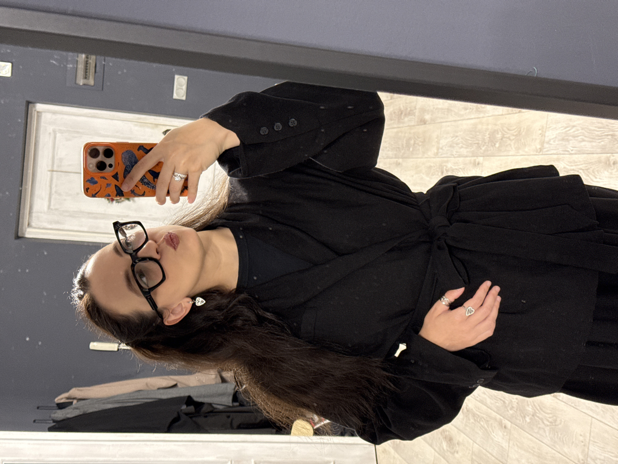 A woman is taking a mirror selfie in an indoor setting. She has long, dark hair, is wearing black-rimmed glasses, and has on a black outfit consisting of a collared jacket tied at the waist and a dark top. She is also wearing heart-shaped earrings, a ring on her left hand, and a ring on her right index finger. She is holding an orange phone with a colorful case in her right hand. The background shows a closet with clothes hanging, a white door, and a grey wall. The floor appears to be made of light-colored wood planks. There is no visible indication of the city of Chaikovsky, Russia.