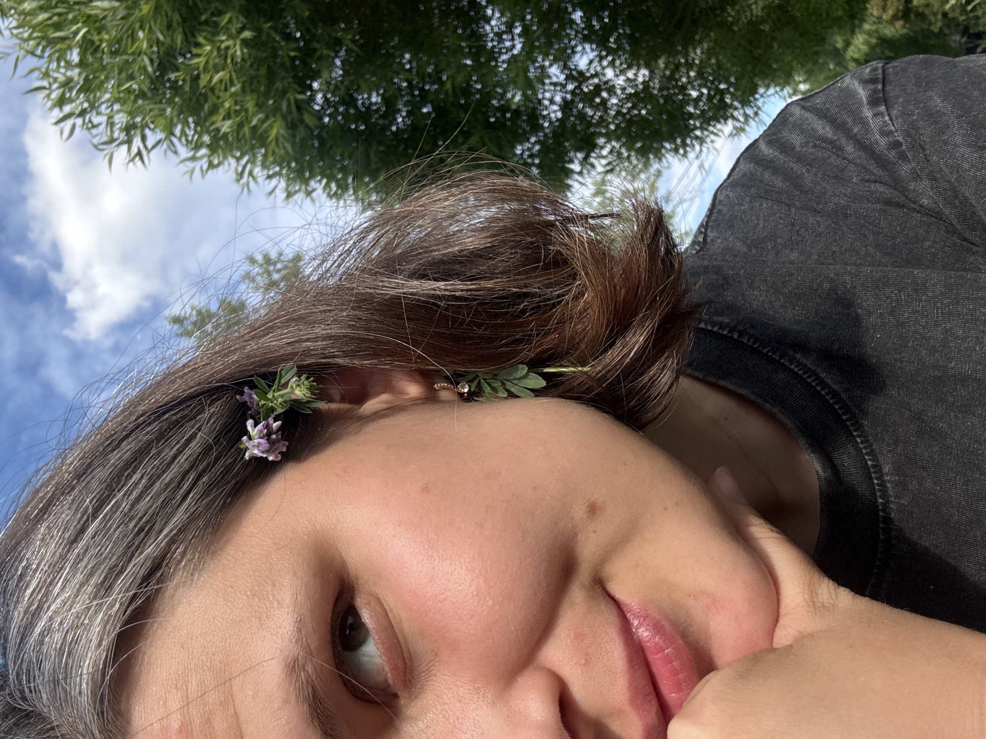The image shows a close-up, profile view of a person's face and shoulder, outdoors. The person has fair skin with some freckles and rosy cheeks. Their left eye is visible, looking upwards and to the left. They have brown hair with streaks of grey, cut to chin length. Small purple flowers are tucked into their hair above their left ear, and a delicate gold earring with green leaves hangs from the same ear. Their lips are slightly parted and appear to be glossy. Their left hand is resting under their chin, with fingers softly curled. They are wearing a dark grey, textured t-shirt. The background consists of a bright blue sky with scattered white clouds and blurred green foliage from trees. The overall impression is one of a casual, natural setting, possibly a park or garden. There is no indication of the city of Chaikovsky, Russia in the image.