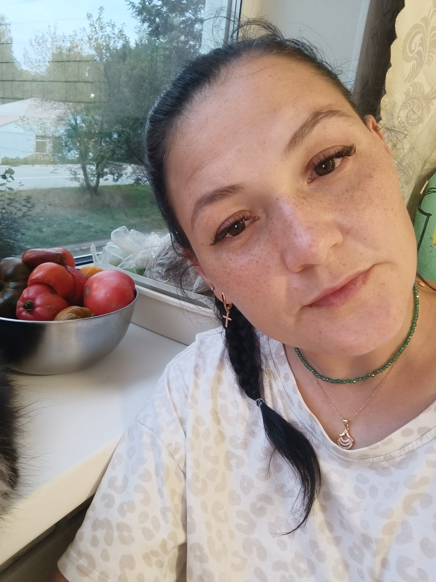 A close-up, slightly angled selfie shows a woman with dark hair, pulled back into a braid, and freckled skin. She is wearing a white t-shirt with a light tan leopard print pattern, a green beaded necklace, and a gold pendant necklace. She has on small gold cross earrings. To her left, in the background, is a bowl of tomatoes and other produce on a windowsill. Through the window, trees and a building are visible, suggesting an outdoor or semi-outdoor setting, possibly in Chaikovsky, Russia, given the location of the original image. The lighting suggests it is daytime.