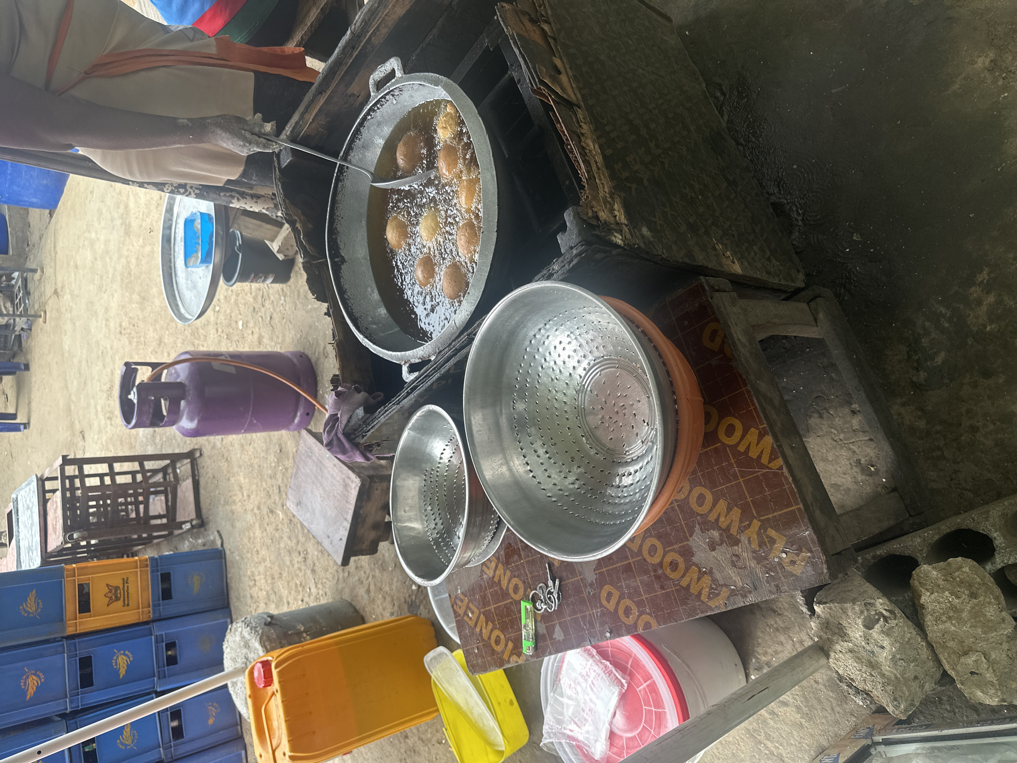 A street food vendor is frying spherical dough-based food items (possibly puff-puffs) in a large metal wok filled with hot oil. The vendor, partially visible, is using a slotted spoon to maneuver the items. The cooking setup consists of a wooden table/stand, a propane tank, and metal colanders for draining. There are blue and yellow plastic crates stacked nearby, and the scene is likely taking place on a street corner in Abuja, Nigeria.