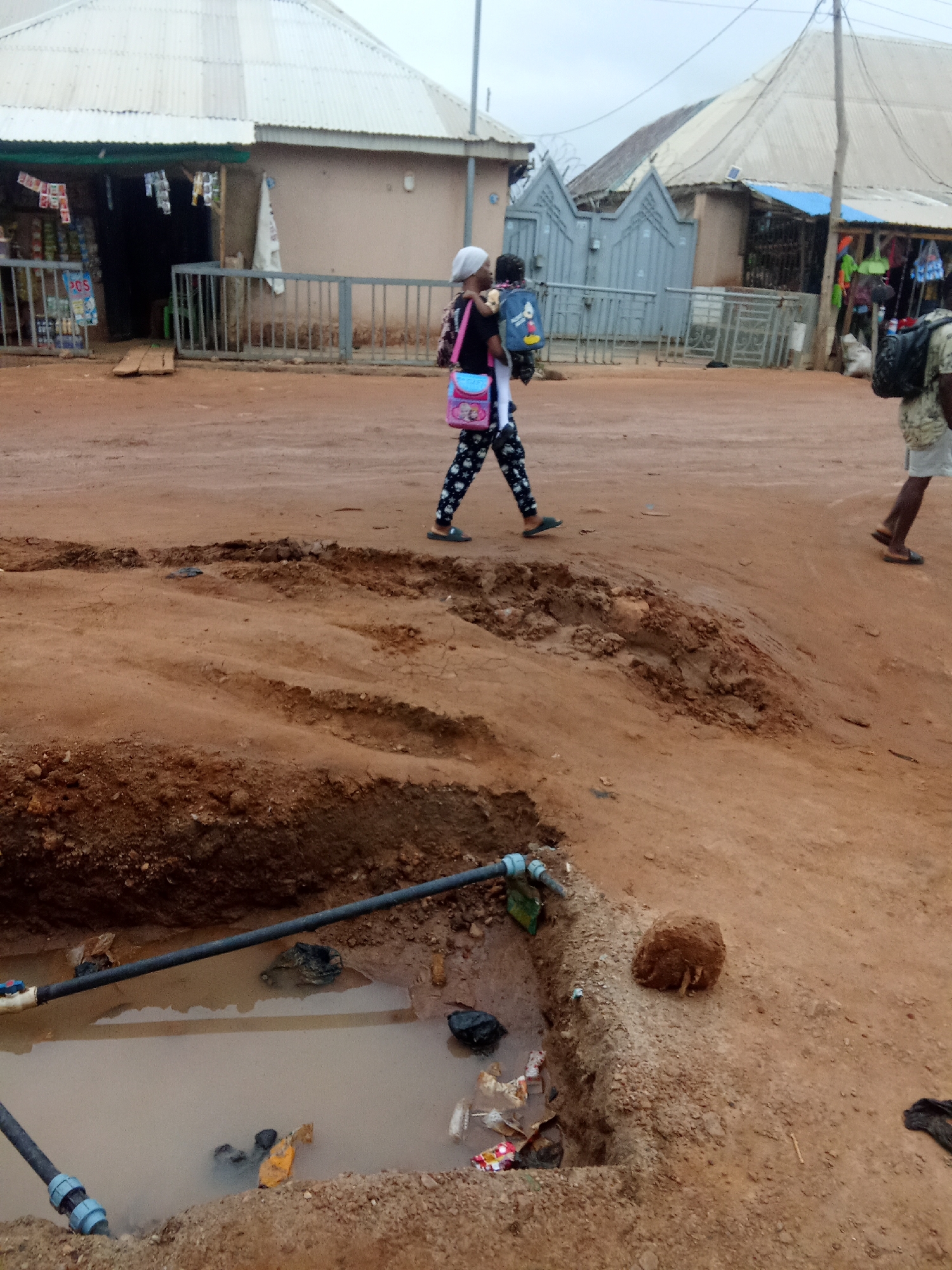 In Kagini, Nigeria, a woman walks carrying a child on her back. She wears a headscarf, black shirt, and patterned pants, with a pink purse slung over her shoulder. The child wears a blue backpack. The ground is a dusty red dirt road, with a large excavation in the foreground containing a muddy puddle and exposed pipes. In the background, corrugated metal roofs of buildings line the street, with some appearing to be small shops. Another person is seen walking away from the camera on the right side of the image, wearing a light-colored shirt and shorts, and carrying a backpack.