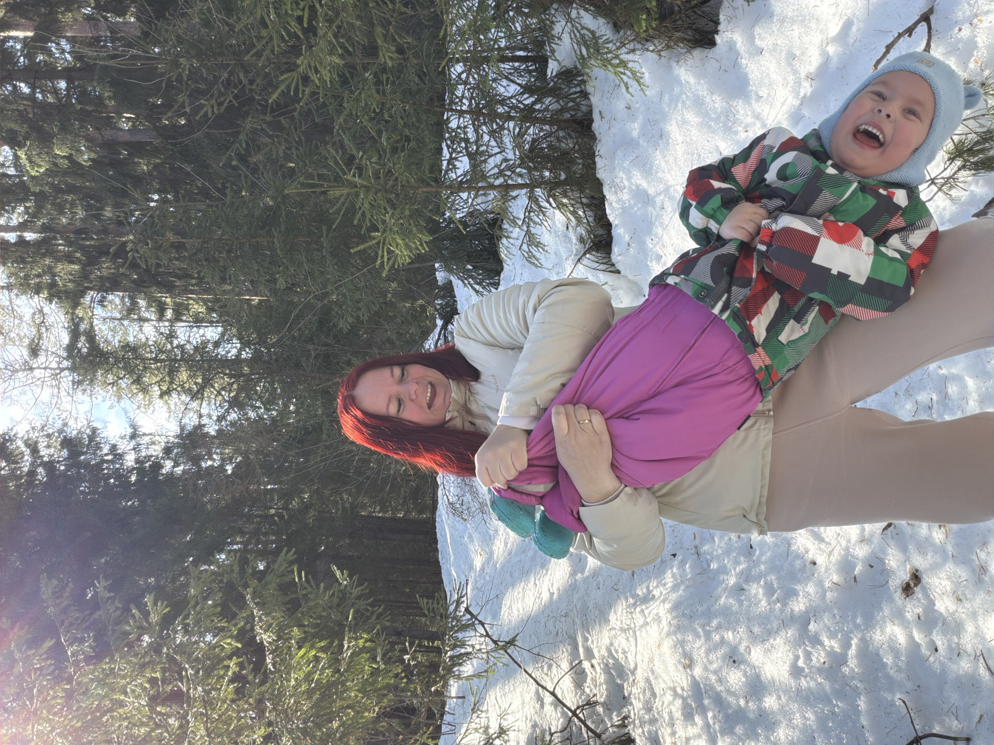 The image displays an adult female holding a young child upside down in a snow-covered forested area. The scene is brightly lit by direct sunlight filtering through the trees. The adult, a female with red hair, is wearing a light beige winter jacket and beige pants; she has a ring on her left ring finger and is smiling, looking down at the child. The child, appearing to be a toddler, is dressed in a multi-colored (red, green, white, black) patterned winter jacket, a bright pink layer, and a light blue knit hat with ear flaps. The child is smiling widely, with an open mouth, positioned with their head towards the bottom of the frame. Both individuals exhibit smiling expressions. The ground is covered in a layer of white snow, showing some disturbances and patches of underlying earth or debris. The background consists of a dense forest primarily composed of tall evergreen trees, with smaller evergreen saplings and some bare branches visible in the mid-ground. The specific location within Chaikovsky, Russia, is identified as a forested park or natural area.