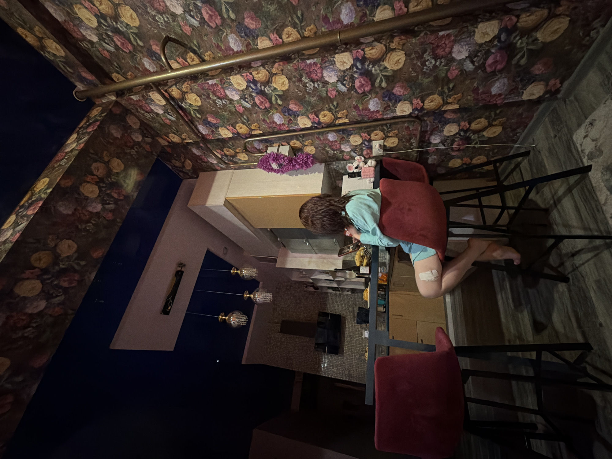 A medium-shot captures an interior scene within an establishment, possibly a restaurant, in Sochi, Russia. A female figure, situated at a counter or table, is seen engrossed in an activity, possibly reading or using an electronic device. Her attire includes a light blue shirt, and a bandage is visible on her leg. The environment features a patterned wallpaper with floral designs adorning the ceiling and walls. The counter is lined with various objects, including what appears to be food and drink containers. Red chairs are positioned around the table. The image has been rotated, giving a disorienting perspective.