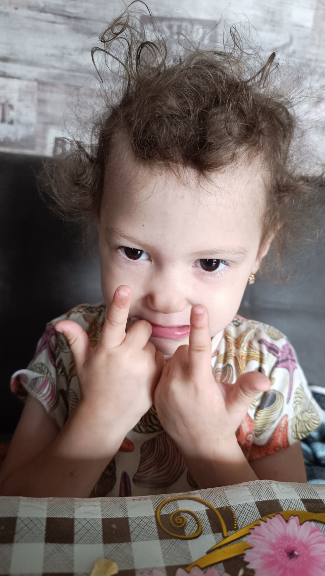 A young girl with messy, curly brown hair is sitting at a table, looking directly at the camera. Her eyes are dark brown, and her lips are slightly parted, with her tongue visible. She is holding her hands up in front of her face, with her fingers pointing towards her cheeks. She is wearing a light-colored t-shirt with a pattern of seashells. The background appears to be a wall with a wood-grain pattern. The table in front of her is covered with a checkered tablecloth. The image does not provide any information about the location in the city of Karsha, Russia.