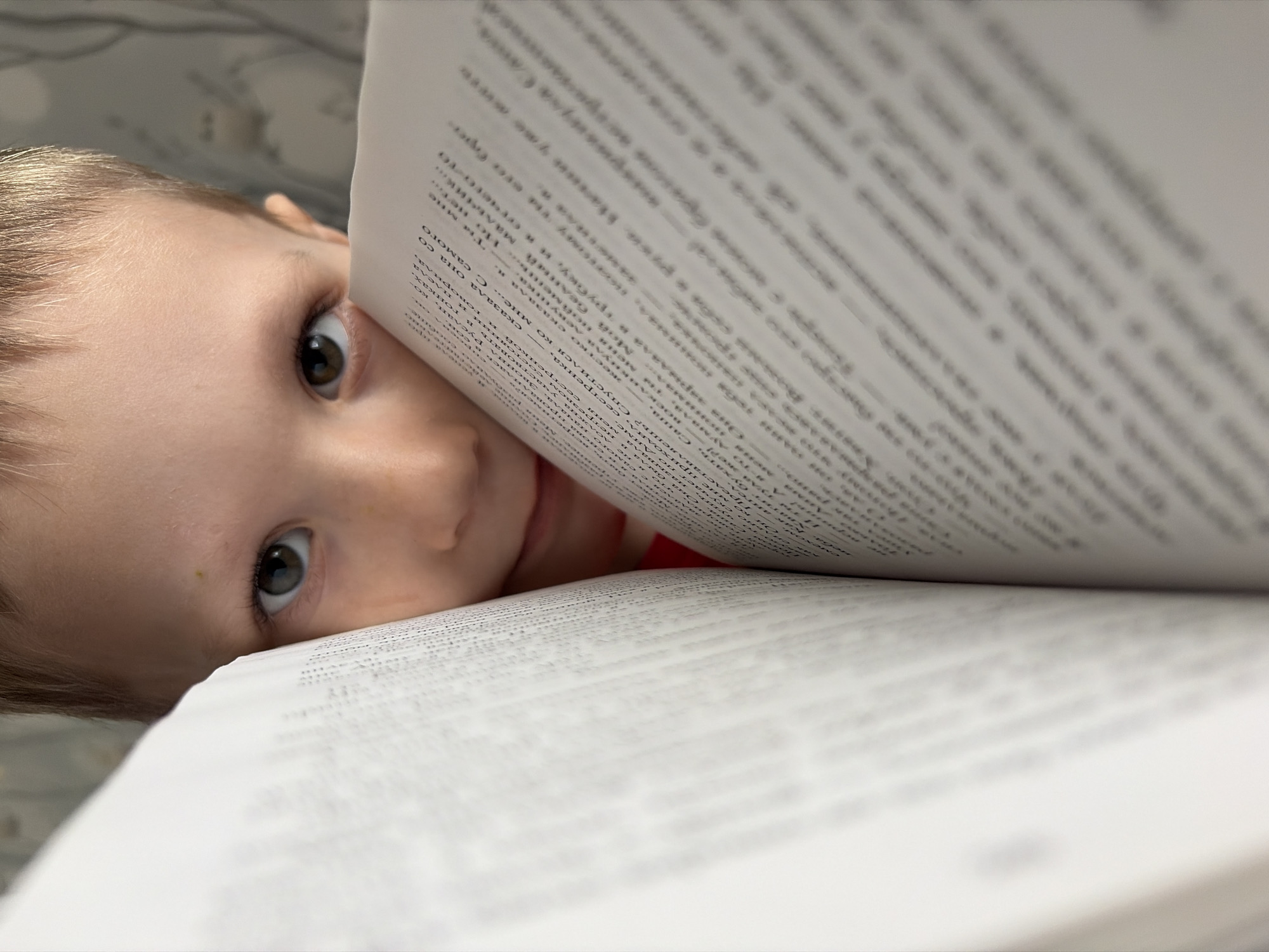 A young child peers over the top of a large, open book, his face angled towards the camera. The book's pages are filled with text in Russian, but the details are blurred due to the shallow depth of field. The child has light skin, brown hair, and large, expressive eyes. A small mole is visible on his forehead. The background is soft and out of focus, possibly suggesting a domestic environment. The location is Chaikovsky, Russia, though this is not directly evident from the image content. The scene suggests a moment of curiosity or playfulness, where the child is engaging with the book in an unconventional way.