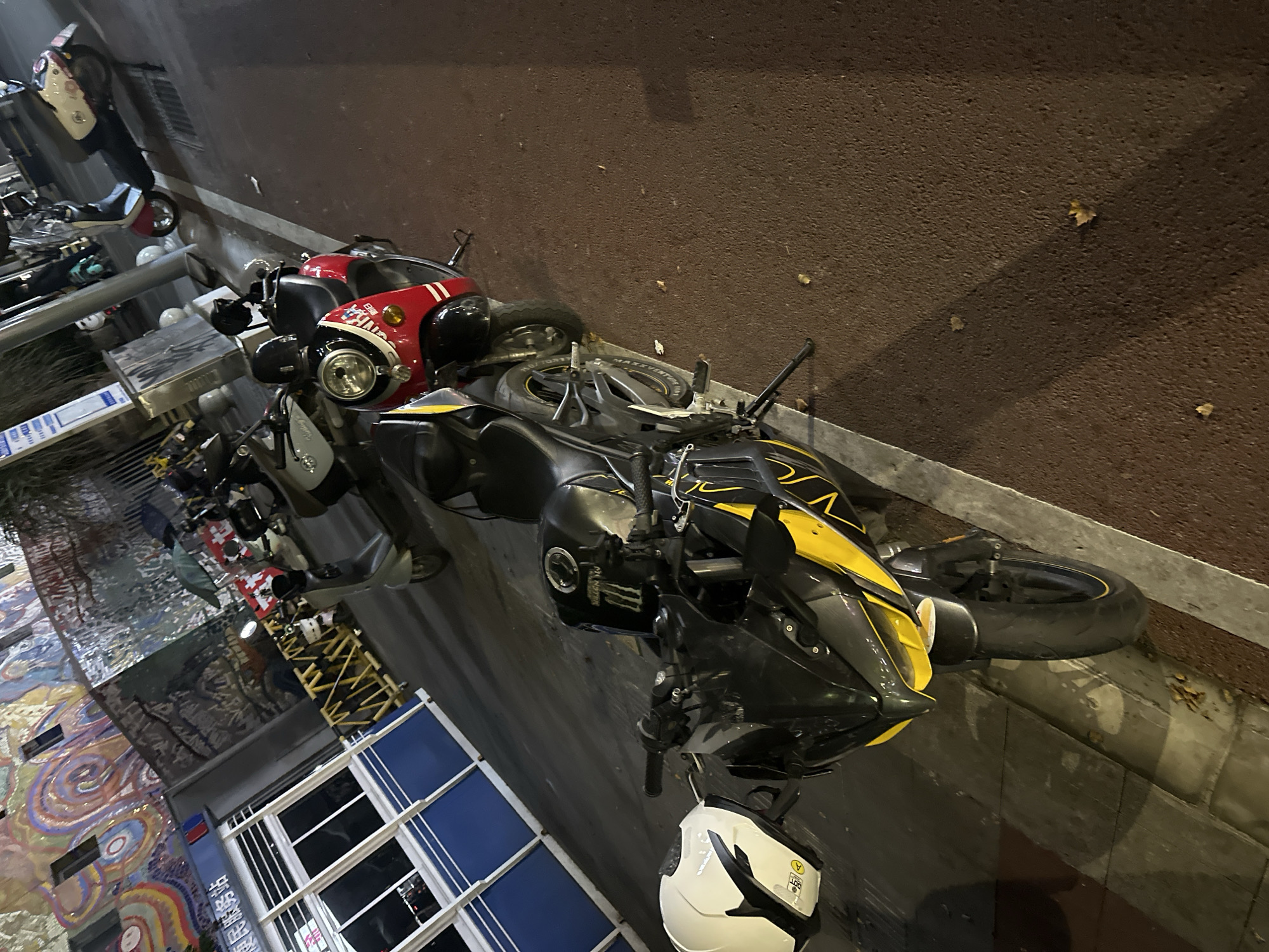 This image captures a street scene in the Chongqing High-tech Zone, China, at night. The foreground features a black and yellow sport motorcycle parked on the side of the road. A white helmet with black accents rests on the motorcycle's handlebars. Behind this motorcycle, a red and black scooter is parked, followed by other scooters and motorcycles further down the street. In the background, a building with a colorful mural is visible, along with what appears to be a service station or kiosk. The street is paved with asphalt and bordered by a curb. The lighting suggests it is nighttime, with artificial lights illuminating the scene. There are no people prominently visible in the frame.