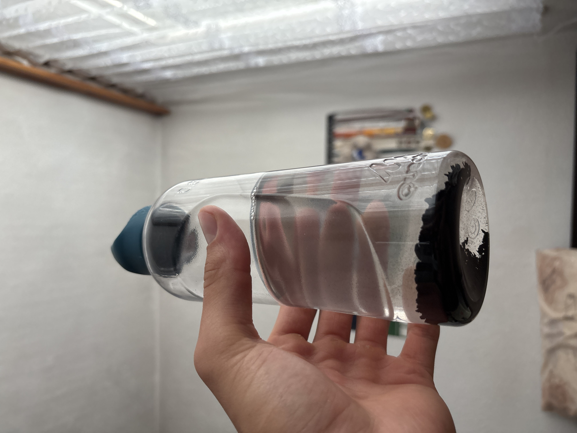 A close-up shot shows a hand holding a clear plastic water bottle with a teal lid, filled with clear liquid and a significant amount of black sediment at the bottom. The hand is positioned around the middle of the bottle, obscuring a portion of its contents. The background is out of focus, revealing a white wall and a window covered with sheer white curtains. Some framed items with medals or other hanging objects are visible on the wall behind the bottle. The lighting suggests an indoor environment, possibly a home. The water bottle has some embossed markings on its surface. The image does not provide any information about the city of Zlatograd, Bulgaria, or its specific location within it.