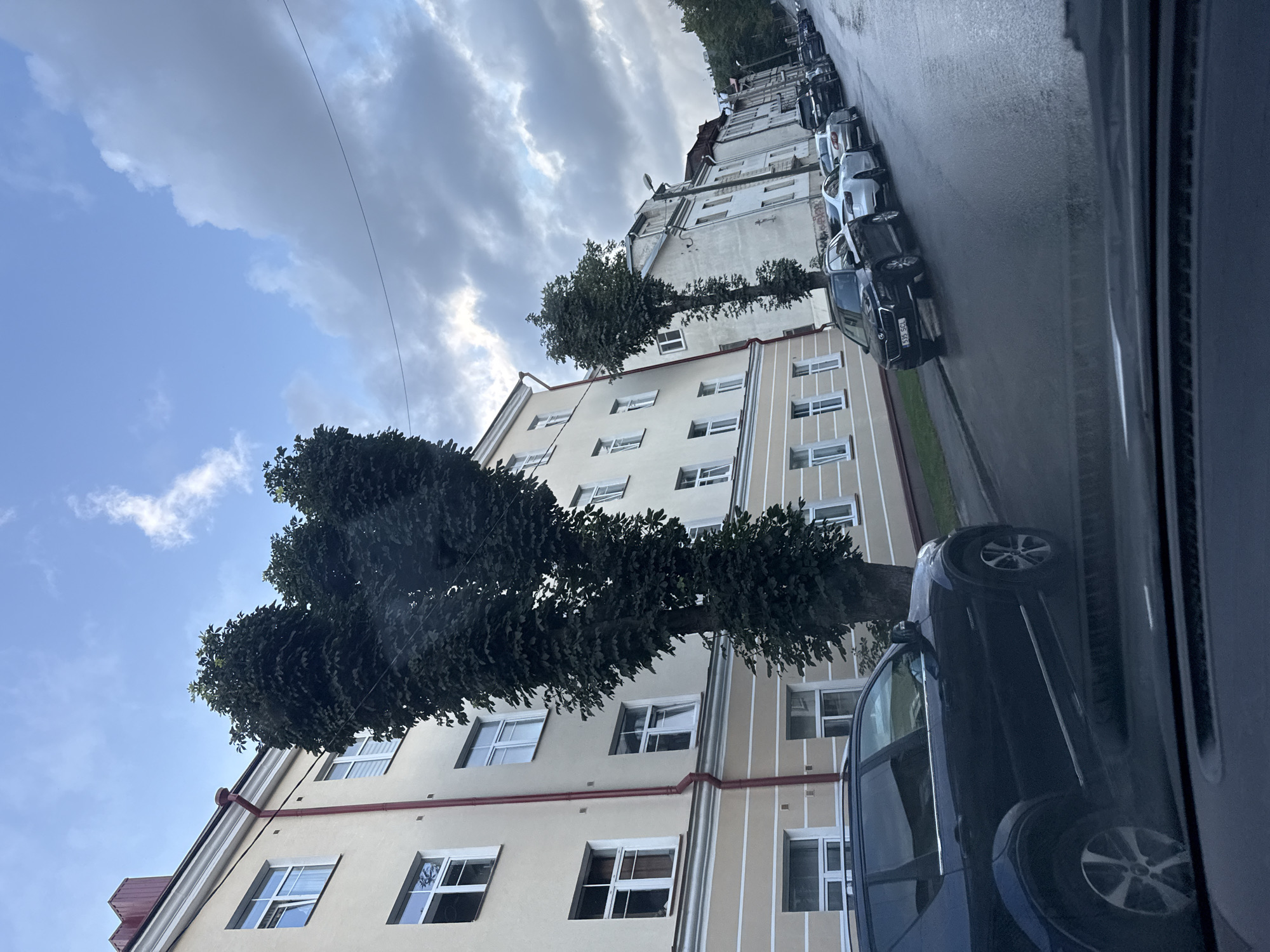 The image shows a street scene in Tallinn, Estonia. In the foreground, a dark blue SUV is parked on the left side of the street. Beyond the SUV, several other cars are parked along the street, including a white sedan and a black sedan. The buildings lining the street are multi-story, with light-colored facades and numerous windows. Tall trees with dense foliage are positioned in front of some of the buildings. The sky is partly cloudy, with a mix of blue sky and grey clouds. The road appears to be wet, suggesting recent rain. The overall impression is of a residential or commercial street in an urban environment.