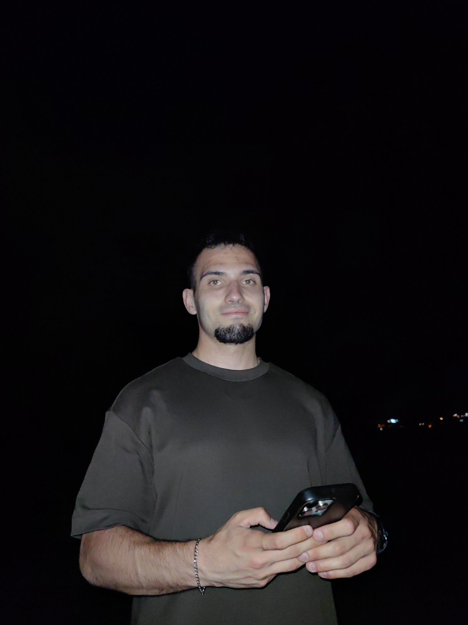 A medium shot depicts a man holding a phone in his hands, set against a dark background. He is wearing a short-sleeved dark grey shirt and has a short beard. The man is also wearing a bracelet and a watch. A few lights are visible in the distance. The location is Unknown, Unknown.