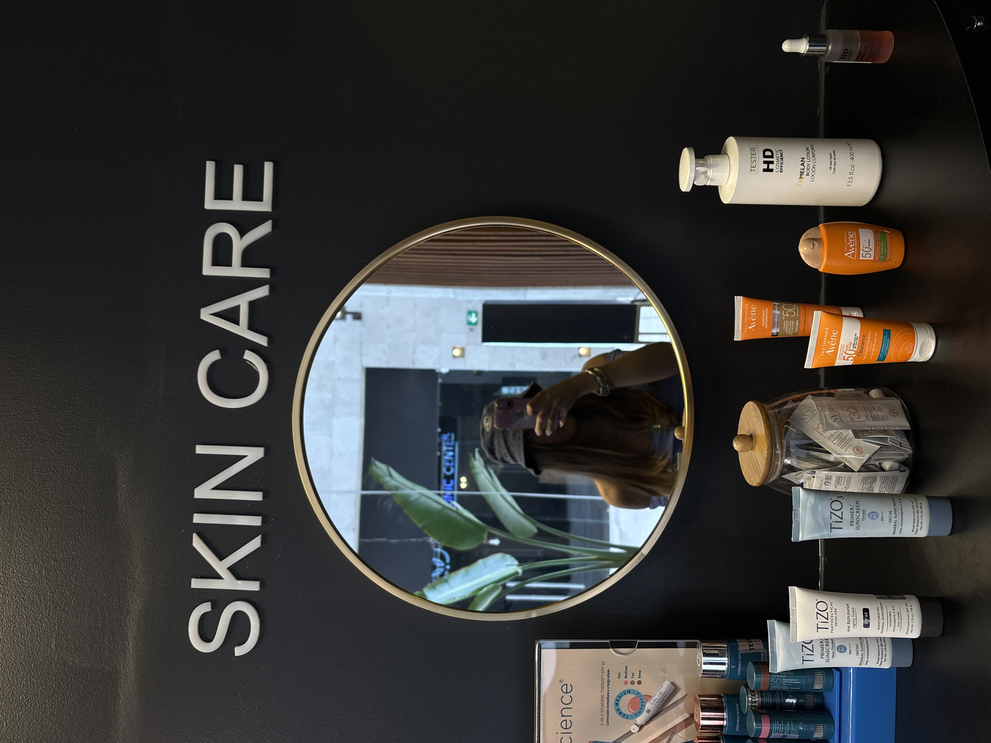 A vertically-oriented photograph shows a display of skincare products, with a reflective, circular mirror in the center. To the left of the mirror, large white lettering spells out "SKIN CARE" vertically. Various skincare items are arranged to the right of the mirror including tubes and bottles of sunscreen and lotions. Brand names such as "Avène" and "TiZO" are visible on the products. There is a jar filled with small packets, possibly samples, and a color cosmetics display tray at the bottom right. The mirror reflects the photographer holding a cell phone, a potted plant, and the exterior of a business identified by a sign that includes the words "IC CENTER", which places the scene in Ciudad López Mateos, Mexico.