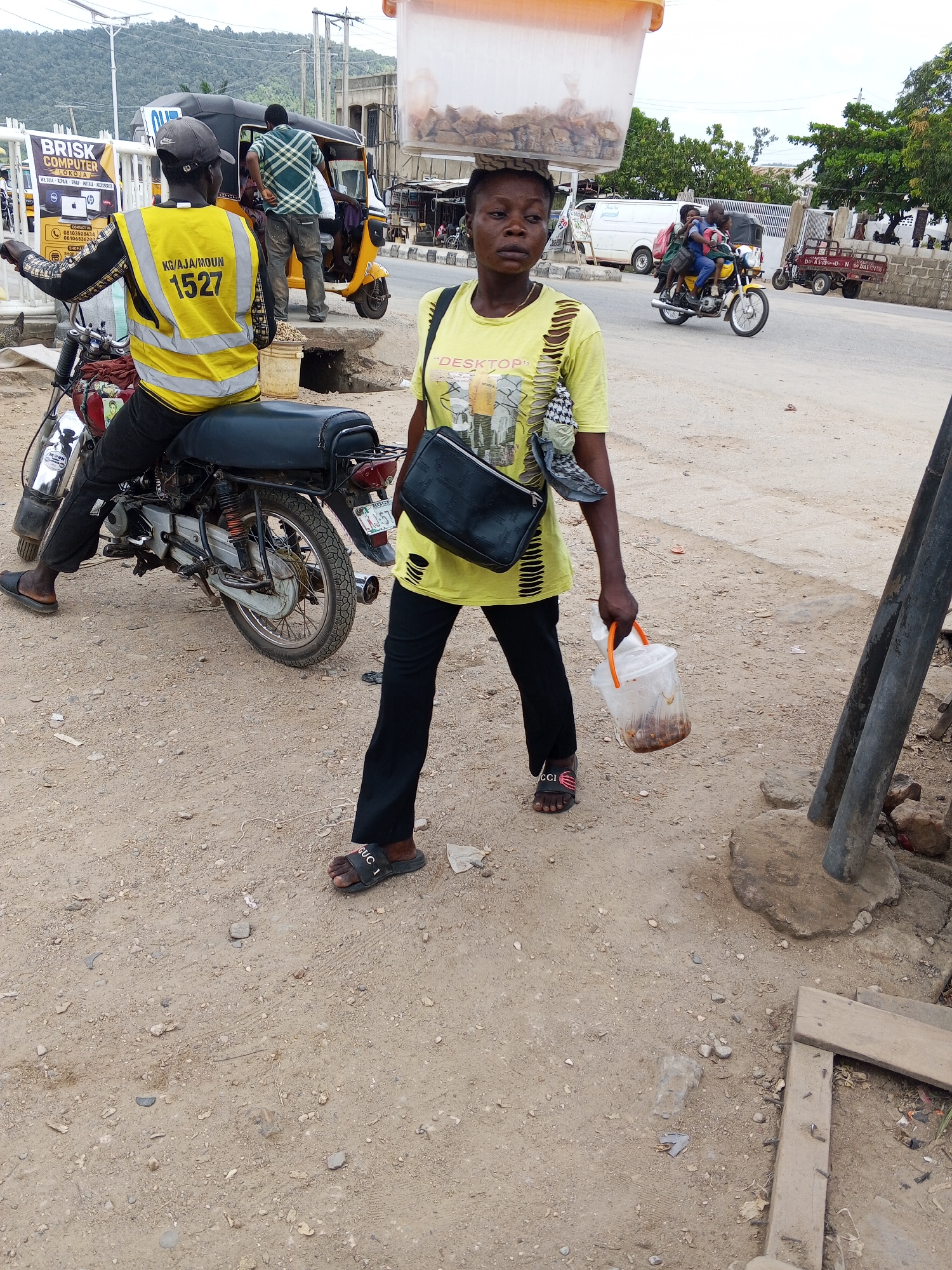 This is a medium shot, eye-level photograph taken outdoors in what appears to be a dusty, roadside environment in Zango-ndaji, Nigeria. The scene is likely urban or peri-urban, with a paved road in the background and a dirt-covered ground in the foreground. The lighting suggests it is daytime, possibly late morning or afternoon, with diffuse sunlight and no harsh shadows, indicating an overcast or hazy sky.
The primary subjects are two individuals. In the foreground, a woman wearing a yellow t-shirt with torn sleeves and black trousers is walking towards the right of the frame. She has a black handbag slung over her shoulder and is carrying a clear plastic bucket with an orange lid in her right hand, containing some sort of food item. She has a container of food balanced on her head. She appears to be looking slightly to her left, with a neutral or perhaps weary expression. Her black flip-flops have "GUCCI" written on them.
To the left of the woman, a man is seated on a motorcycle. He is wearing a yellow reflective vest over a black and white patterned long-sleeved shirt, and black trousers. The vest has the text "KG/AJA/MOUN 1527" on the back. He is looking towards the left of the frame, seemingly engaged in conversation or observing something off-camera. The motorcycle is a common model, painted red and black.
In the background, a yellow auto-rickshaw (keke) is visible, with at least two people inside. Further in the background, a white van and a truck with a flatbed are parked. A sign for "BRISK COMPUTER" is prominently displayed on a white structure to the left, with contact numbers and text in English. The overall impression is one of everyday life and commerce on a busy Nigerian street.