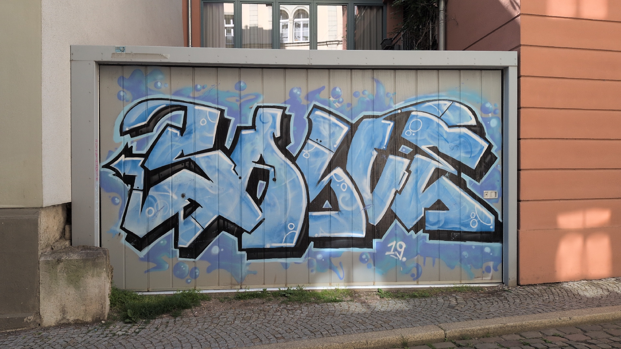 The image depicts a section of a garage door in Weimar, Germany, adorned with blue and black graffiti. The graffiti features stylized, blocky letters with shading and bubble-like elements. To the right of the garage door is a salmon-colored building. The ground in front of the garage is paved with cobblestones and has a curb. A small patch of grass and weeds grows at the base of the garage. In the upper portion of the image, reflections of a building with multiple windows are visible on the garage door. A small, light-colored sticker is affixed to the metal frame above the graffiti.