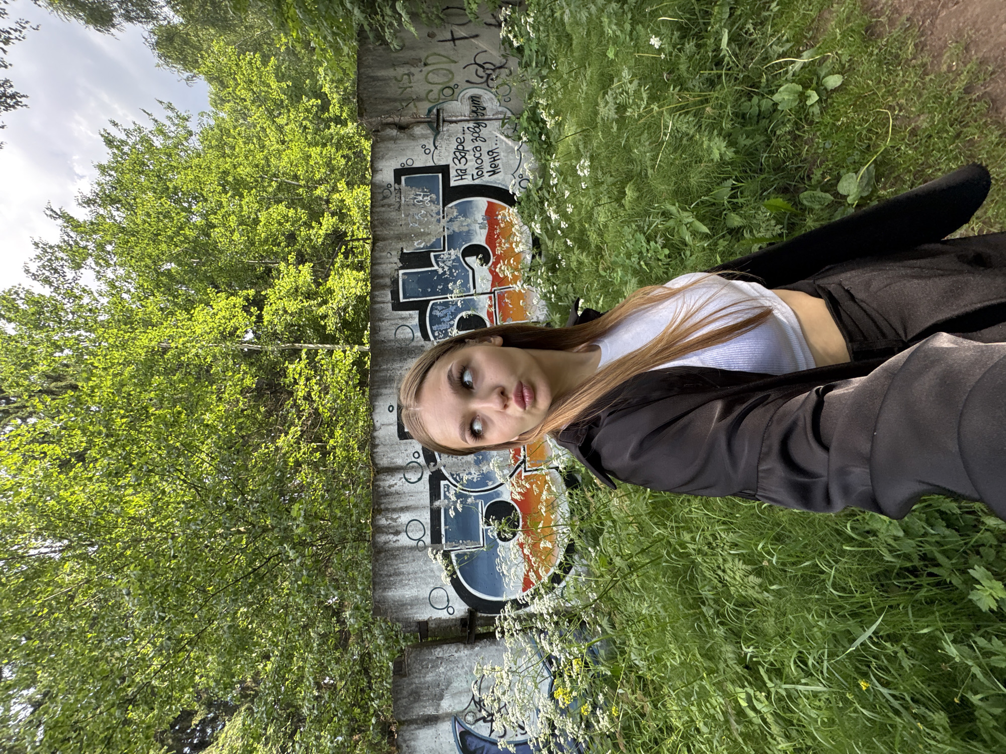 A young woman with long, blonde hair is pictured from the chest up, holding a camera to take a selfie. She is wearing a white crop top and a black open jacket. Her lips are pursed as if she is about to kiss the camera. She is standing in front of a graffiti-covered concrete wall, surrounded by lush green vegetation and some wildflowers. The wall appears to be part of an abandoned structure, possibly an old industrial building, which are common in Chaikovsky, Russia, a city known for its industrial history and presence of birch forests. The background is dominated by the dense green foliage of trees, suggesting a natural or semi-natural environment within or on the outskirts of the city.