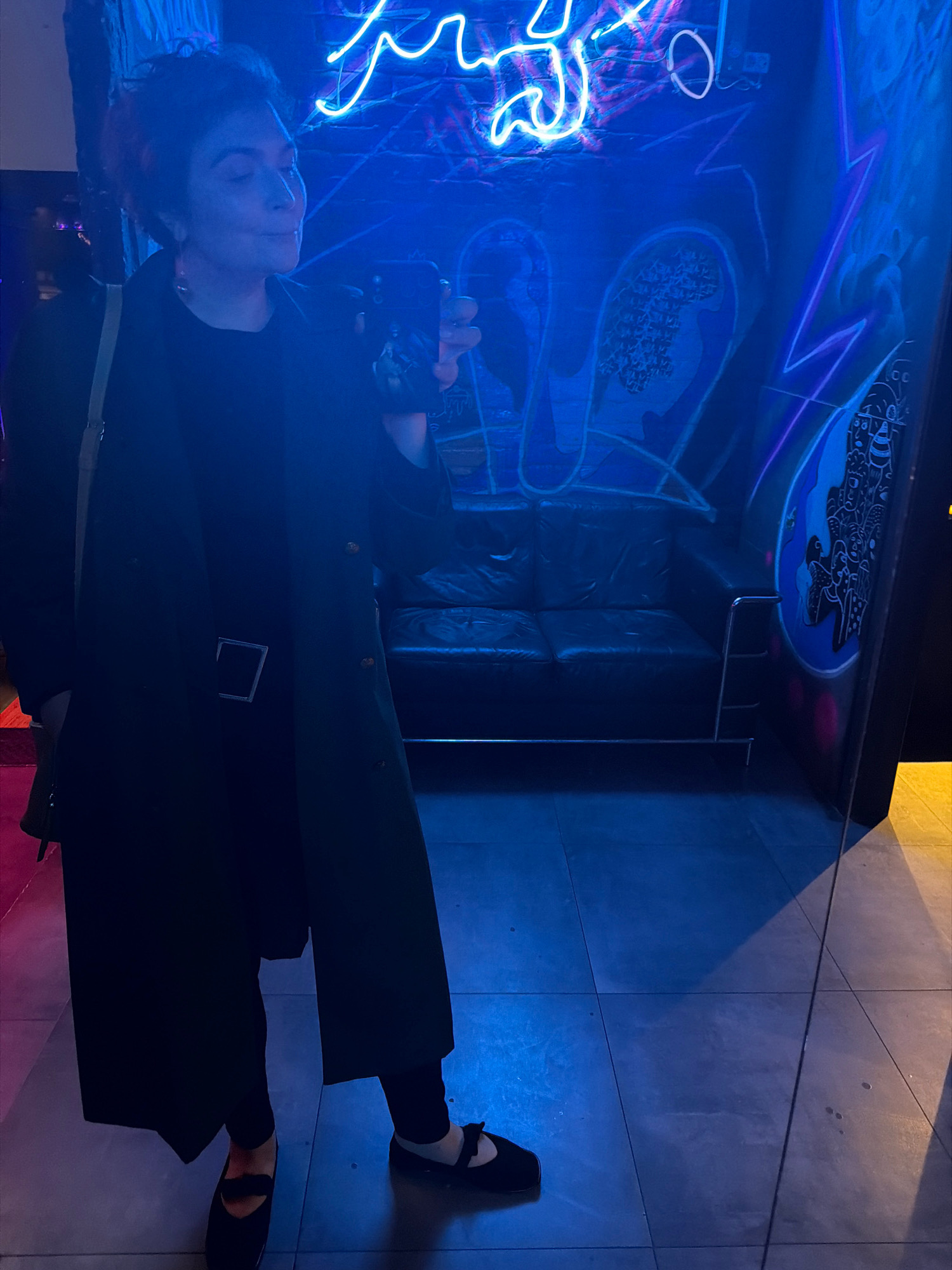 A full-shot, low-angle, vertical mirror selfie shows a person standing in a dimly lit space with blue and pink neon lights. The person is wearing a dark trench coat with a silver belt buckle, dark pants, and black flat shoes with white accents. They are holding a smartphone in their right hand, taking the picture. The background features a black leather couch against a wall adorned with graffiti and bright blue and pink neon lights. The floor is tiled and reflects the ambient light. The overall impression is of a trendy, urban environment. This scene does not provide enough information to determine its location in Samara, Russia.