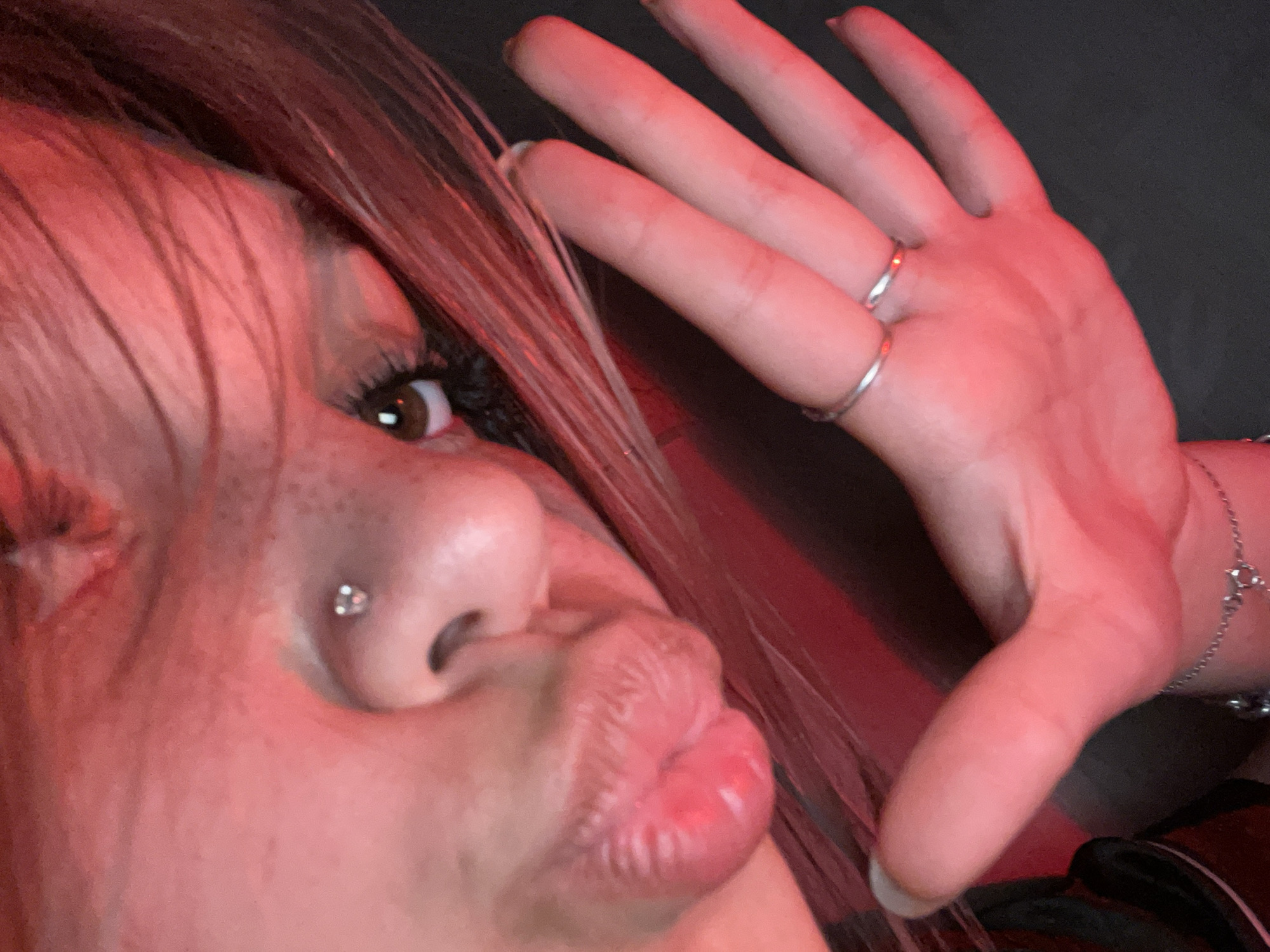 The image is a close-up, low-angle shot of a person's face and hand, illuminated by red light. The person has a nose piercing, styled eyelashes, and is puckering their lips. They are wearing two silver rings on their fingers, and a silver bracelet with a heart-shaped charm is visible on their wrist. The background is dark and appears to be an indoor setting. There is no information in the image to suggest a location in Saint Petersburg, Russia.