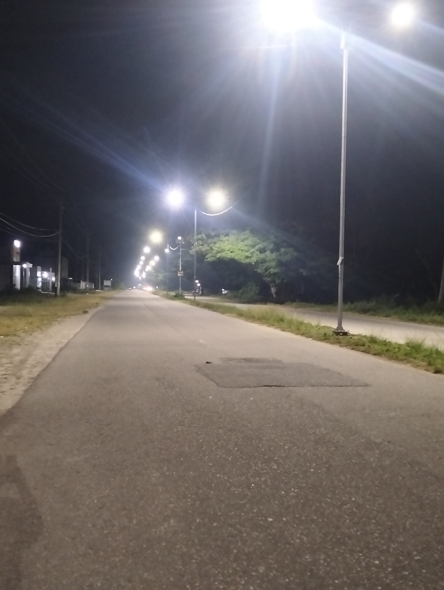 This is a low-angle, eye-level, full shot of a paved road at night, illuminated by streetlights. The road is lined with grass and trees on both sides. There are several streetlights visible, some of which are very bright and create lens flares in the image. There are also some buildings and power lines visible on the left side of the road. In the distance, there appear to be some lights from vehicles. The image appears to have been taken in a somewhat rural or suburban area, possibly on the outskirts of Kampal, Indonesia.