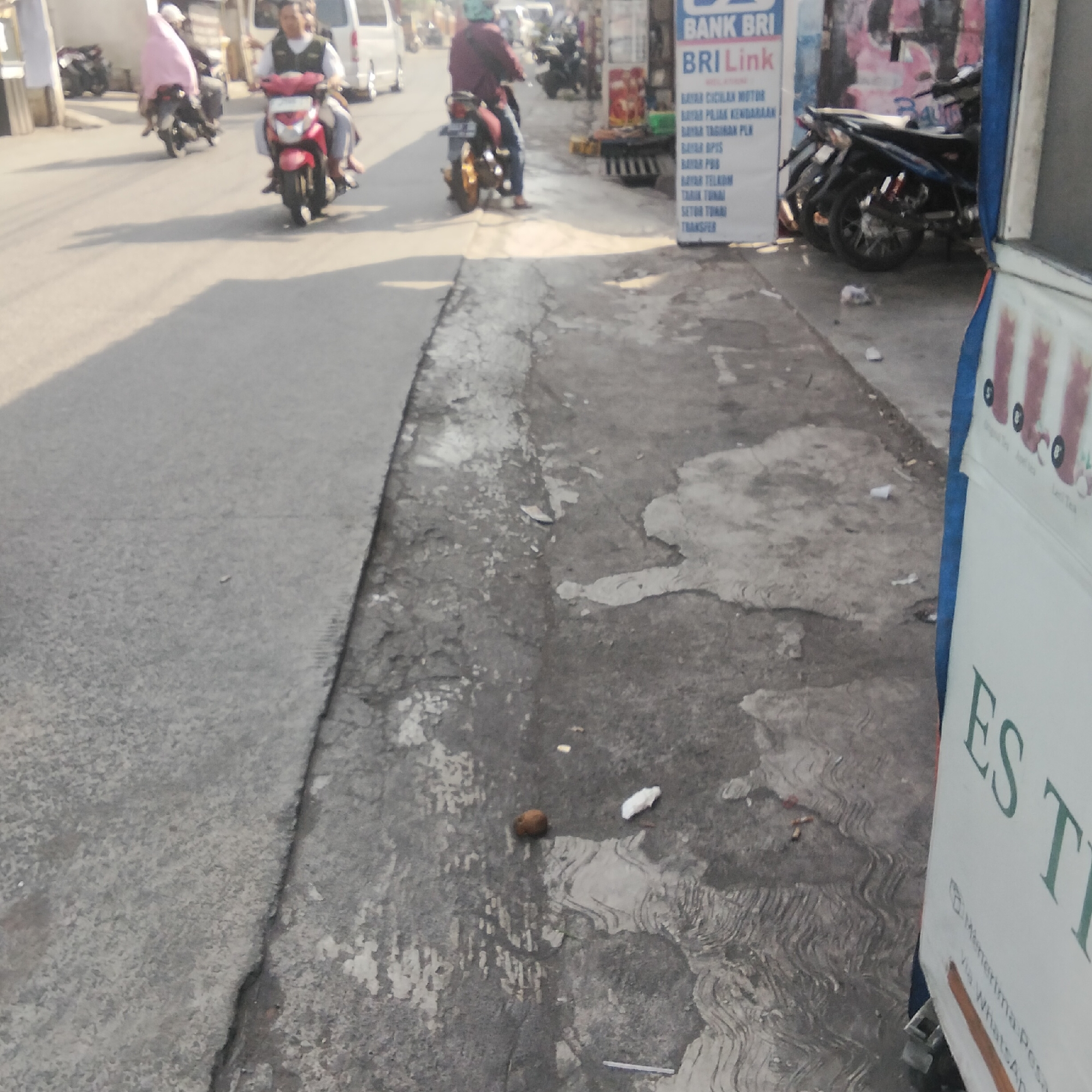 The image captures a street scene in Sukasari, Indonesia, with a focus on the road and sidewalk. Several motorcycles are present, some with riders. In the foreground, the textured concrete sidewalk is cracked and weathered. A small, round object, possibly a piece of fruit or debris, rests on the sidewalk near some scattered white paper scraps. To the right, a portion of a white sign is visible, with green lettering that appears to be advertising something, possibly "ES T". The sign also shows images of drinks. The background shows more motorcycles, a white van, and some buildings. The lighting suggests it is daytime, with shadows cast on the road.