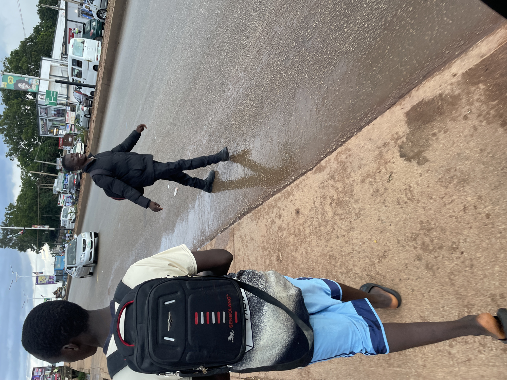 The image shows a view of a street in Jos, Nigeria, with two young men walking along the side of the road. The man in the foreground, facing away from the camera, is wearing a light-colored t-shirt, blue shorts, and black flip-flops, and carries a black backpack. The man in the background, facing the camera, is wearing a black puffer jacket, black trousers, and black shoes, and also appears to be carrying a bag. The road is wet, indicating recent rain. Several vehicles are visible on the road, including a silver sedan, a white van, and other cars. Buildings and trees line the street, with advertising billboards present. The sky is overcast.