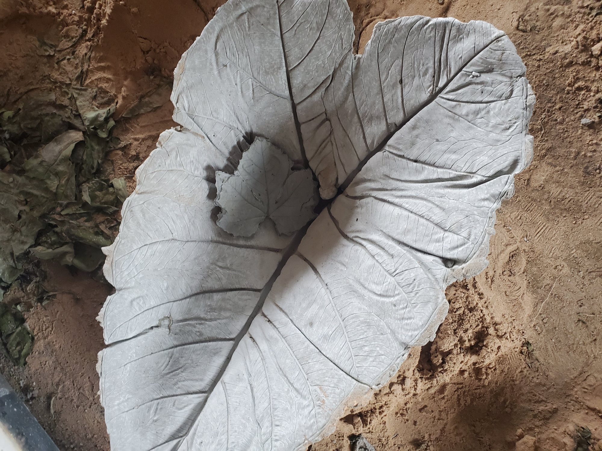 A close-up, overhead shot shows a leaf-shaped concrete casting lying on sandy soil, with some dried leaves visible to the left. The concrete casting, textured to resemble the veins and shape of a large leaf, is the central subject. The sandy ground appears disturbed, possibly from recent activity. The scene is located in the city of Jos, Nigeria.