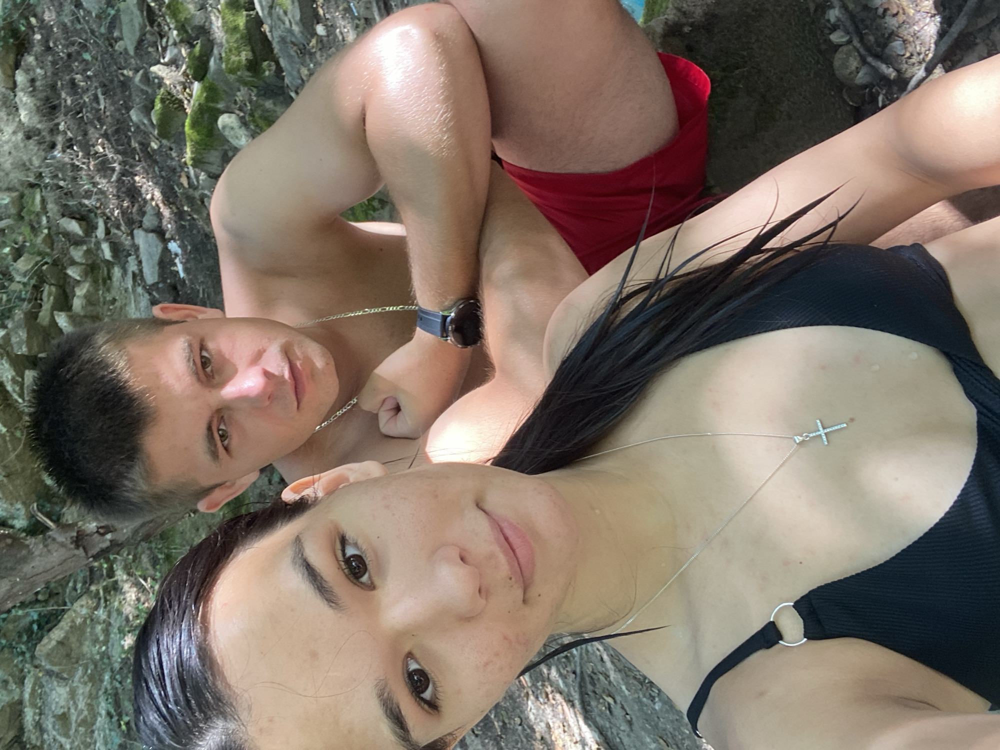 The image shows a young couple in an outdoor setting, likely near a body of water, given the wet hair of the woman and the attire of both individuals. The woman, in the foreground, is wearing a black bikini top and a silver necklace with a cross pendant. Her hair is dark and wet, pulled back into a ponytail. The man is in the background, wearing red swim trunks and a silver chain necklace. He has short, light brown hair and appears to be looking towards the camera. They are seated on rocks in a wooded area. The ground is covered in stones, dirt, and some mossy rocks, suggesting a natural, possibly rural environment. The lighting suggests it is daytime, with sunlight filtering through the trees. The location is identified as Debnevo, Bulgaria.