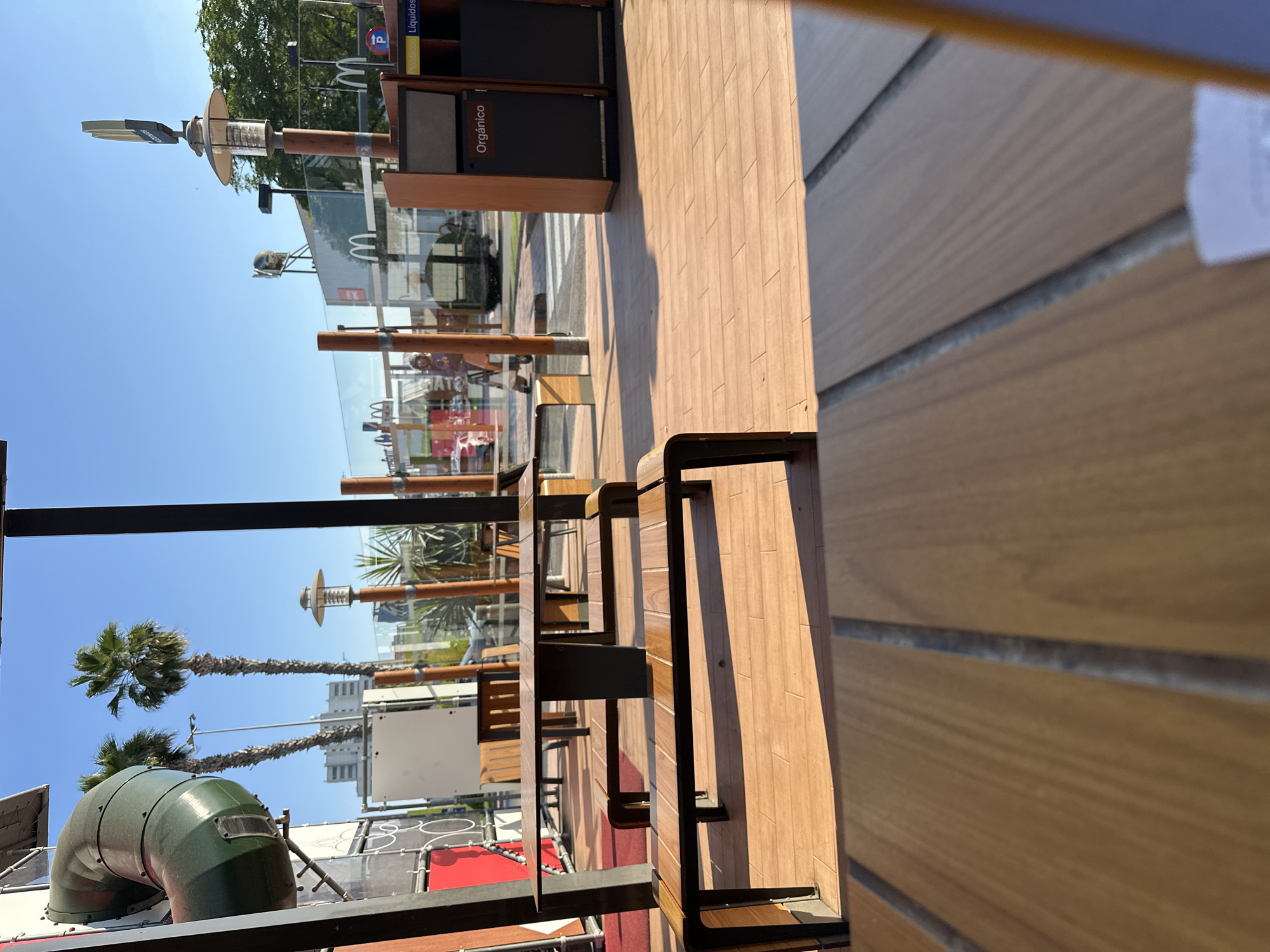 The image shows an outdoor seating area at a McDonald's restaurant in Sedaví, Spain. The scene is captured during daylight with a clear blue sky. In the foreground, wooden picnic tables and benches are visible, with the floor made of wooden planks. Behind the seating area, a play structure with a green tube slide is prominent, along with palm trees and modern buildings in the background. A recycling station with labels for "Orgánico" (organic) and "Plástico/Líquidos" (plastic/liquids) is also present. The environment appears clean and well-maintained, characteristic of a fast-food establishment.