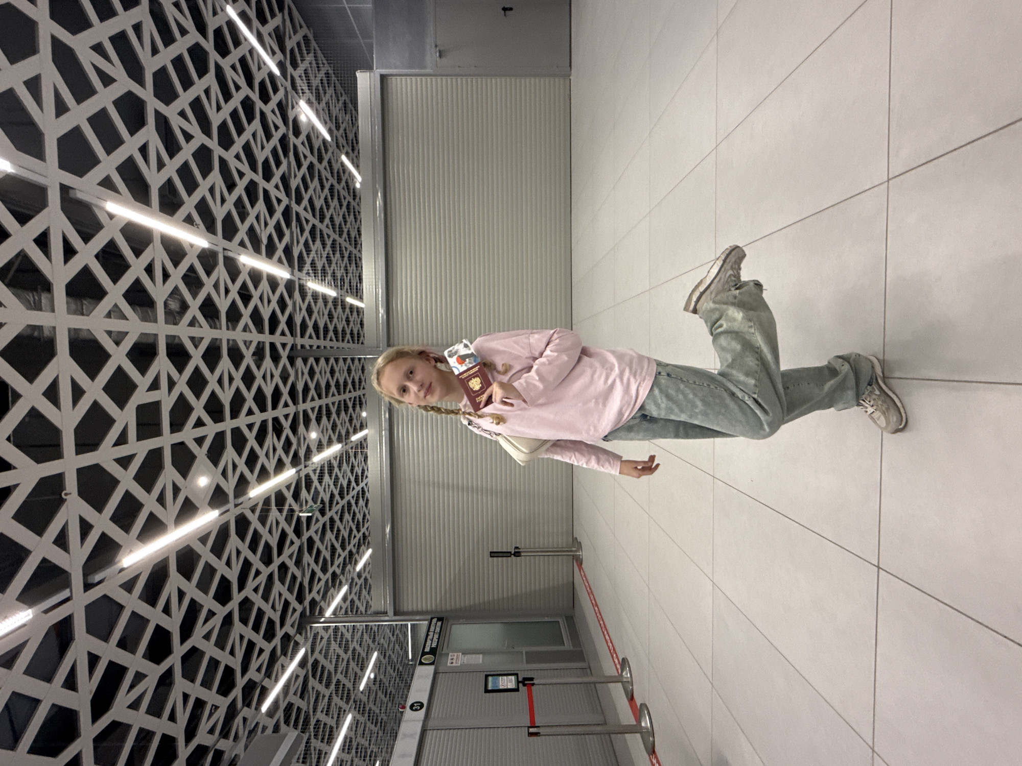 A young girl with blonde pigtails stands in what appears to be a modern airport or transit station, holding a Russian passport. She is wearing a light pink long-sleeved shirt, light blue jeans, and white sneakers. A white purse hangs over her shoulder. The background features a decorative wall with a geometric pattern and embedded strip lights, as well as a roll-down door and a restricted area demarcated by metal stanchions and a red barrier. A sign with the number 31 is visible near the restricted area. The floor is tiled in large, light grey squares. The location is the Ufa International Airport in Russia.