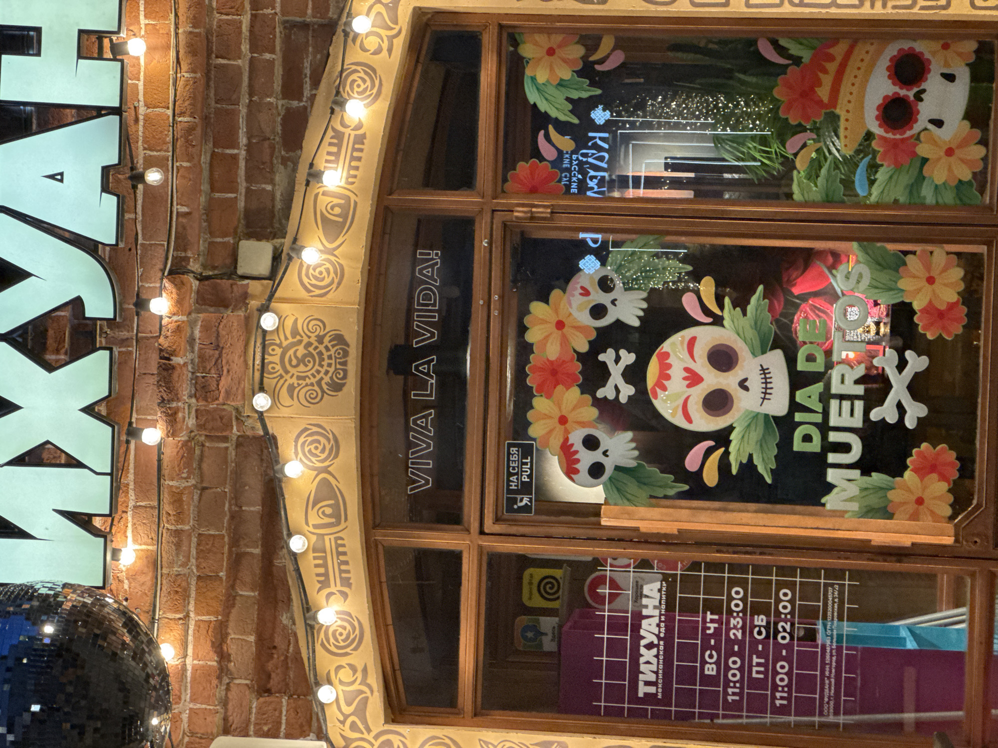 The image shows the entrance to a establishment named "ТИХУАНА" (Tikhuanа) in Nizhny Novgorod, Russia. The establishment appears to be a Mexican restaurant, indicated by the text "МЕКСИКАНСКАЯ ЕДА И НАПИТКИ" (Mexican food and drinks) and the Dia de Muertos (Day of the Dead) themed decorations.
The decorations include stylized skulls (calaveras), marigolds, and green leaves, creating a festive and vibrant atmosphere. The entrance is framed by a yellow facade with ornamental patterns and string lights. Above the entrance, large, light-blue letters spell out part of the establishment's name.
A sign on the left door displays the opening hours:
- VS - ChT (presumably Sunday - Thursday): 11:00 - 23:00
- PT - CB (presumably Friday - Saturday): 11:00 - 02:00
Below the hours, there is a legal notice with company information: "ООО 'ФРУДЭНИК' ИНН: 5260487853 ОГРН: 1225200049707 603000, г. Нижний Новгород, ул. Белинского, Покровская, д. 34/2" (LLC "FRUDENIK", TIN: 5260487853, OGRN: 1225200049707, 603000, Nizhny Novgorod, Belinsky St., Pokrovskaya, bld. 34/2). This confirms the location in Nizhny Novgorod and provides specific street and building details.
The entrance has a door with glass panes, allowing a view into the interior, which is decorated with fairy lights and appears to have plants. A "PULL" sign is visible on the right door, indicating how to open it. The overall scene suggests a lively and culturally inspired dining or entertainment venue.
