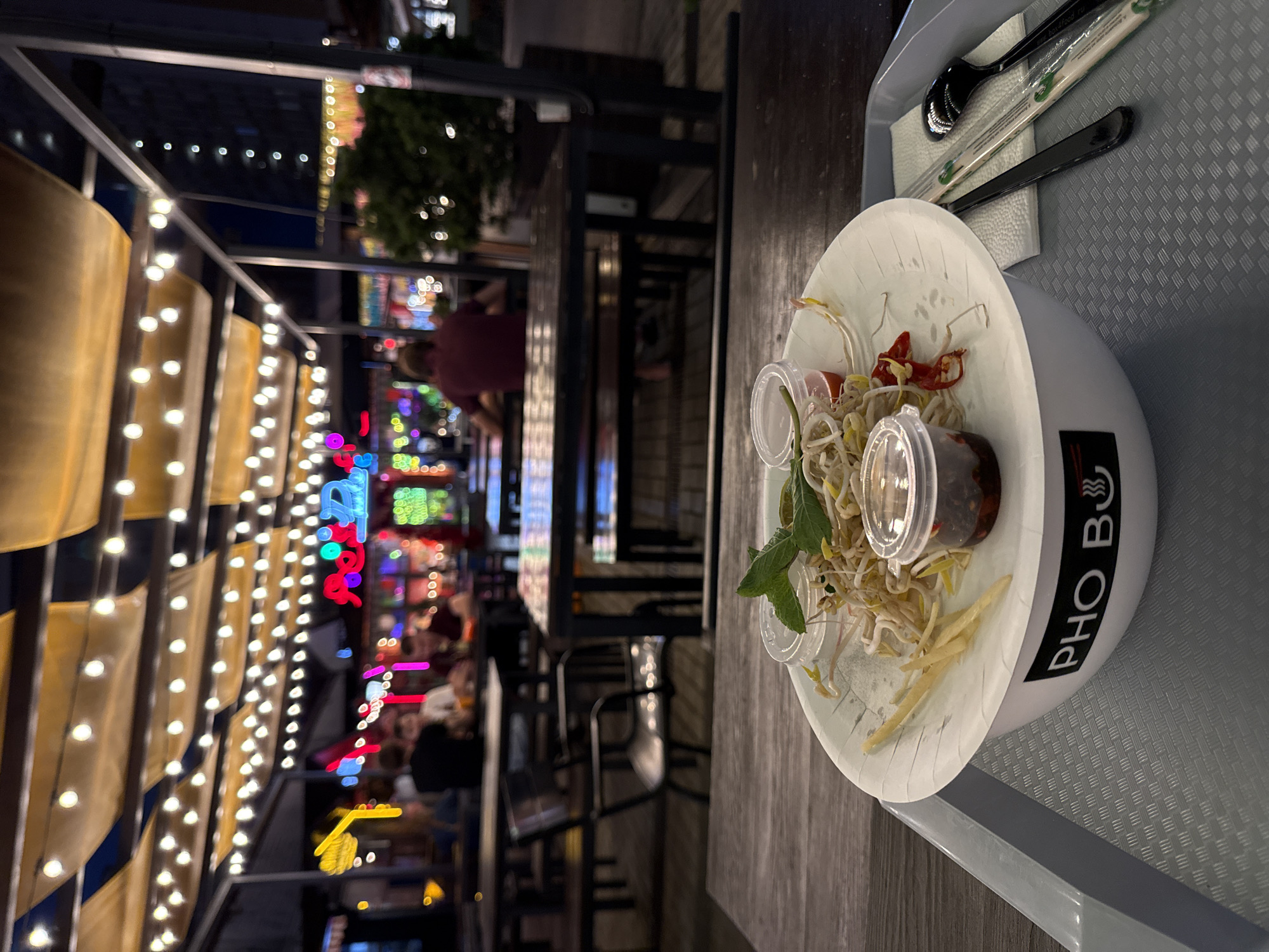 This image shows a close-up of a dish of pho at a restaurant. The pho is served in a white bowl with chopsticks and a spoon resting on top. The bowl has the text "PHO BO" printed on it. The background shows a busy restaurant with people dining and colorful neon lights. The time of day appears to be evening or night. The setting is likely an outdoor seating area given the presence of string lights and the overall ambiance. The overall mood seems lively and bustling.