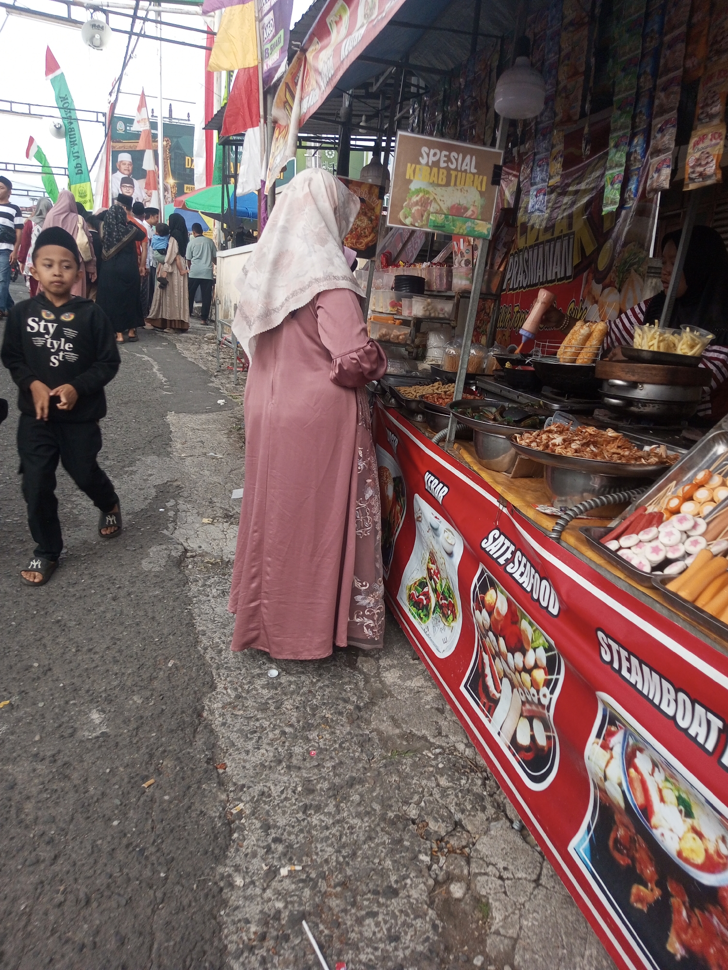 The image captures a street food stall in Pandansari, Indonesia, likely during a daytime event given the festive decorations. The stall, with a prominent red banner featuring images of food and text like "Sate Seafood" and "Steamboat," displays an array of prepared dishes and ingredients. These include skewers of sausage and meatballs, fried food items, and what appears to be a large pan of mussels. A woman in a pink hijab and dress is seen interacting with the stall, and a young boy in a black hoodie and pants walks past in the foreground. Behind the stall, other people and decorations, including flags and banners with Indonesian text, suggest a community gathering or market atmosphere. The ground is paved with asphalt and concrete, showing signs of wear and scattered debris.