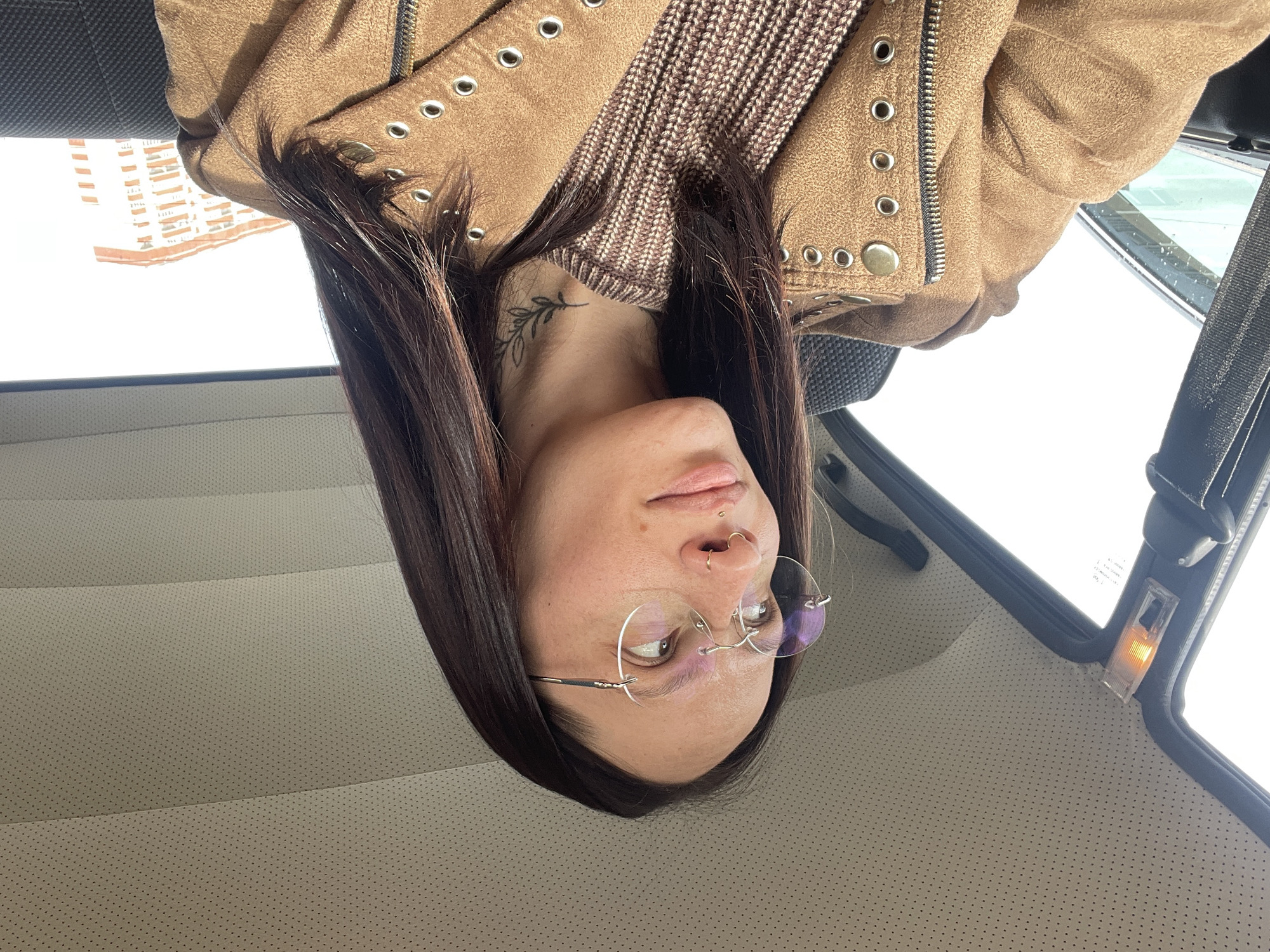 A woman with light skin, brown hair, and glasses is photographed inside a car. She has multiple facial piercings on her nose and is wearing a brown jacket over a brown sweater. A tattoo is visible on her neck. The car's interior features light-colored, perforated upholstery. A small light is on near the ceiling of the car. Through the car window, an apartment building is visible. The location is likely Izhevsk, Russia, based on the context clues provided in the prompt.