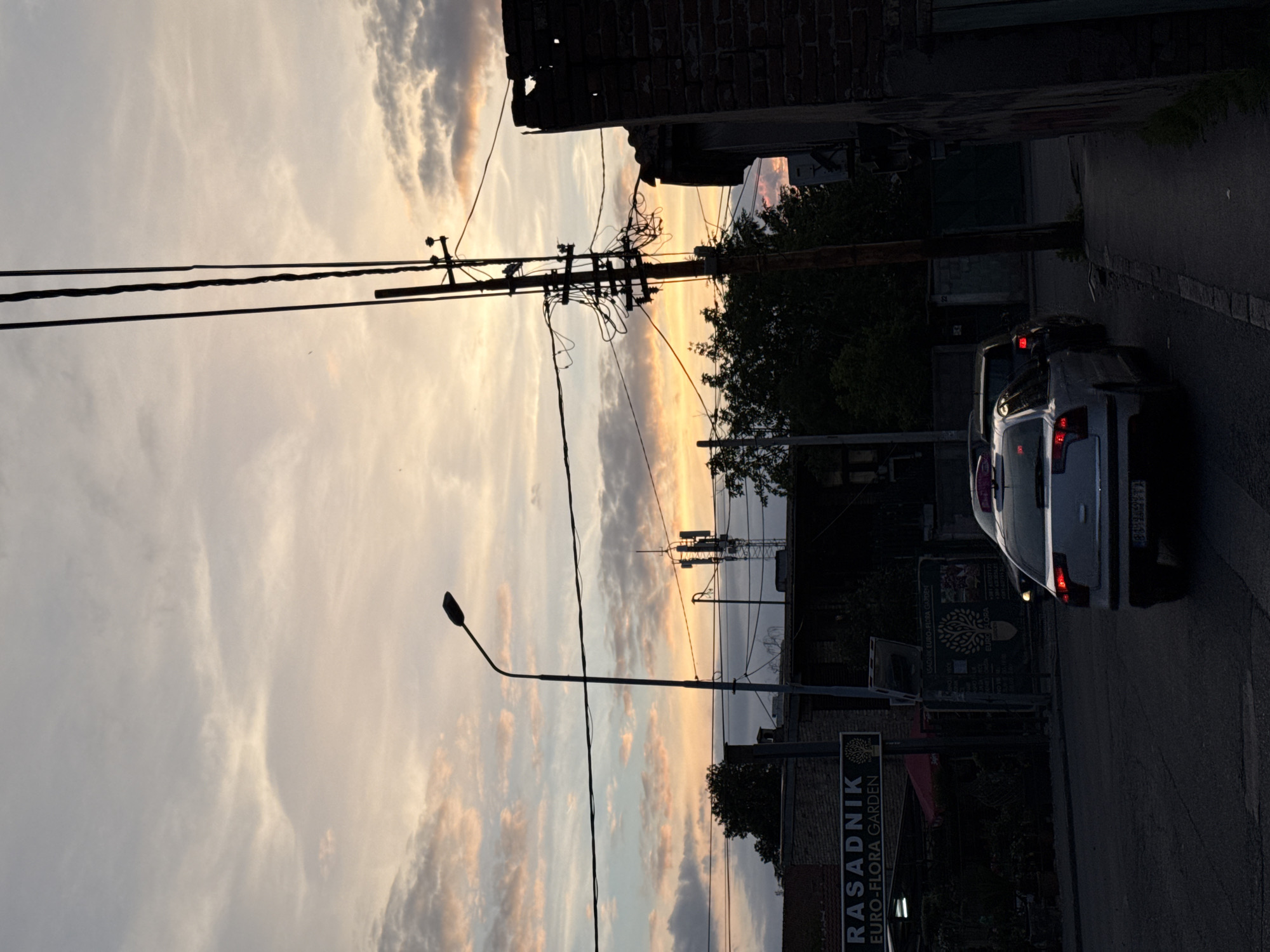 The image depicts a street in Vozdovac Urban Municipality, Serbia, during sunset. The sky is filled with clouds illuminated by the setting sun, casting a warm glow. Overhead power lines crisscross the scene. On the right side of the street, a parked white car is visible, its taillights faintly lit. A building with a brick facade is seen on the left, along with signage that reads "RASADNIK EURO-FLORA GARDEN". A streetlight stands nearby. In the background, a communication tower is visible. The scene appears peaceful.