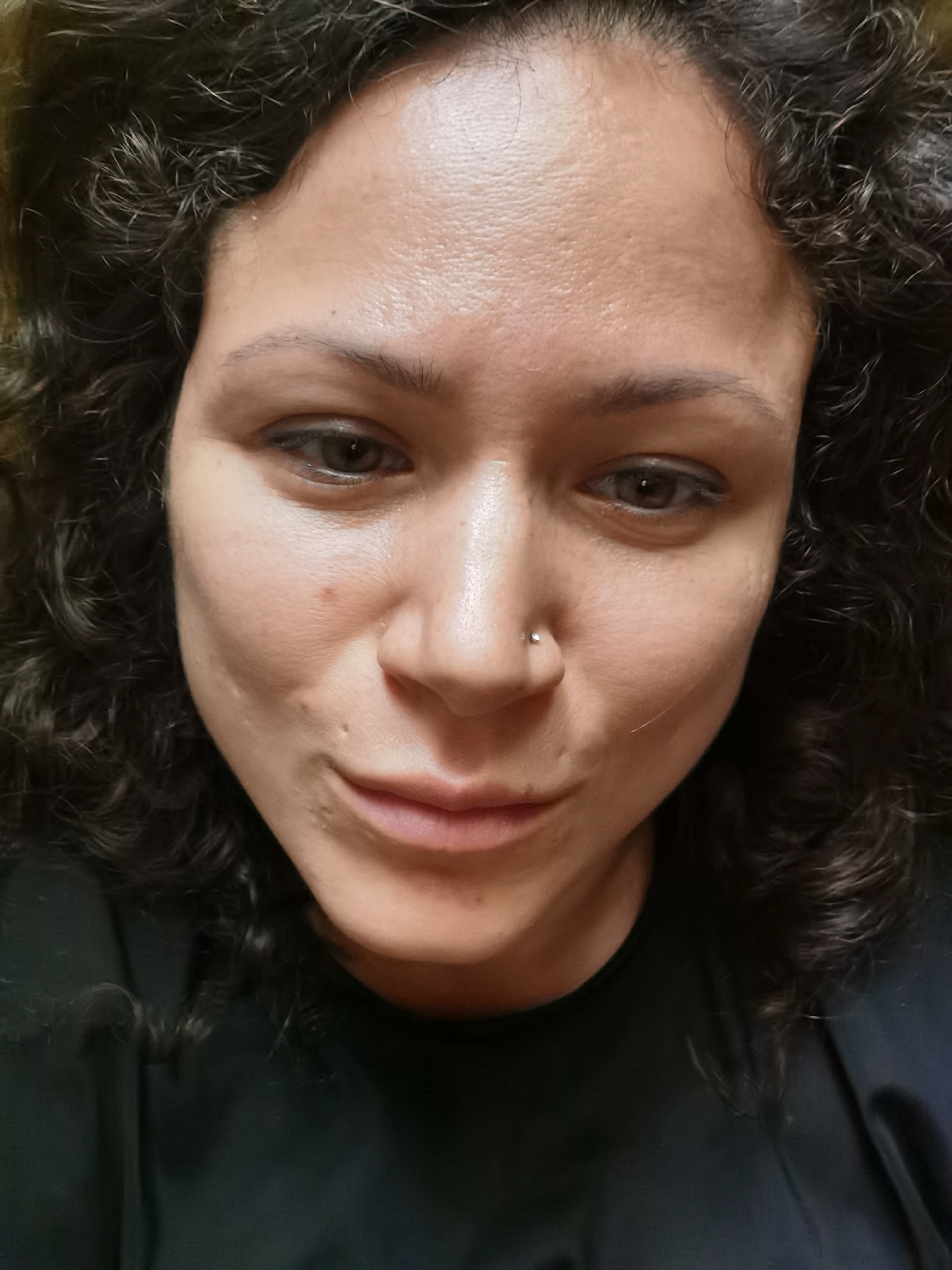 The image is a close-up, head-and-shoulders portrait of a woman with dark, curly hair. She is looking downwards, and her face is slightly tilted. She has a nose ring on her left nostril. The lighting suggests she is indoors. There is no discernible urban environment or reference to Chaikovsky, Russia.